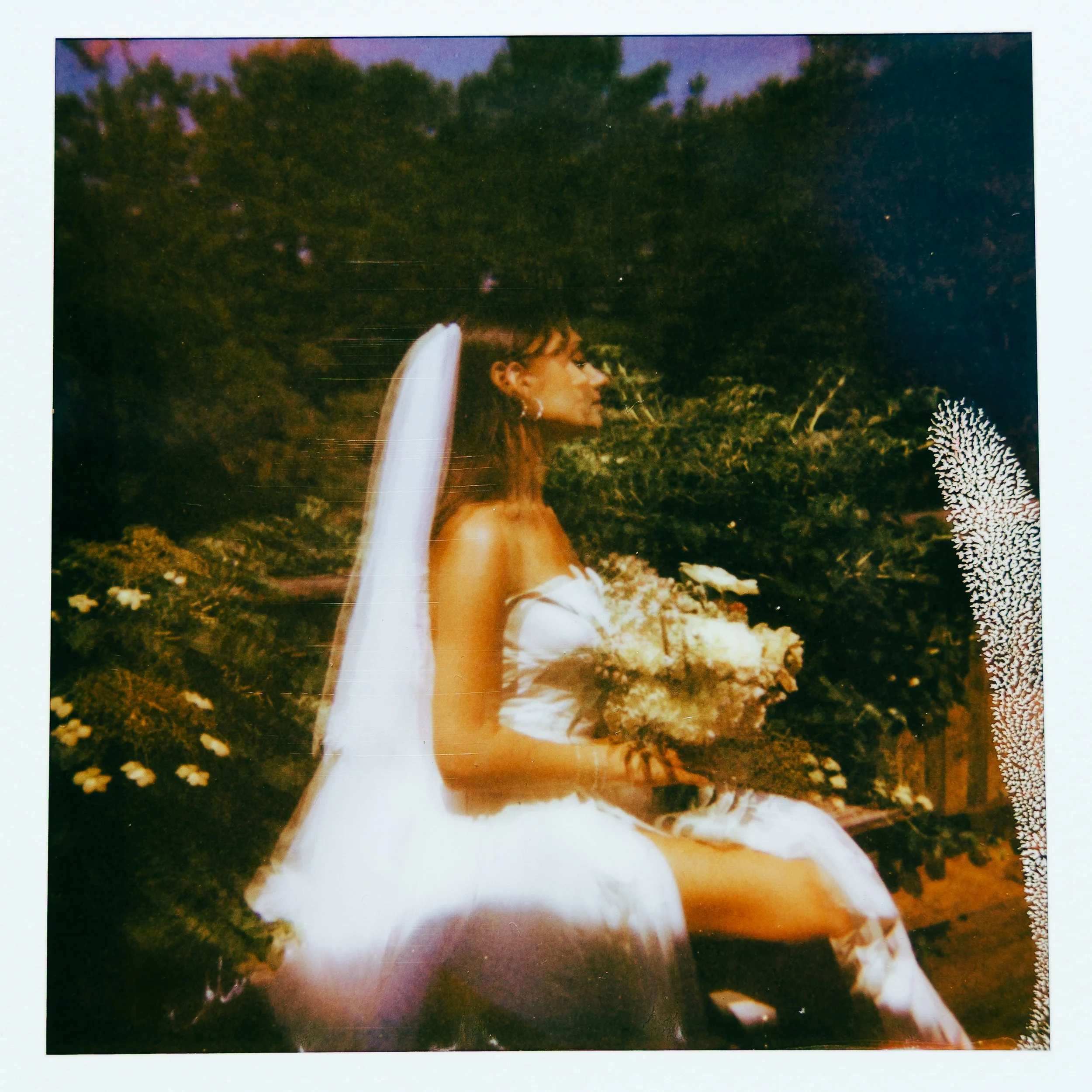 A woman in a white dress with floral details holding a bouquet of flowers, standing outdoors in a garden or park with trees and foliage in the background during dusk or evening.