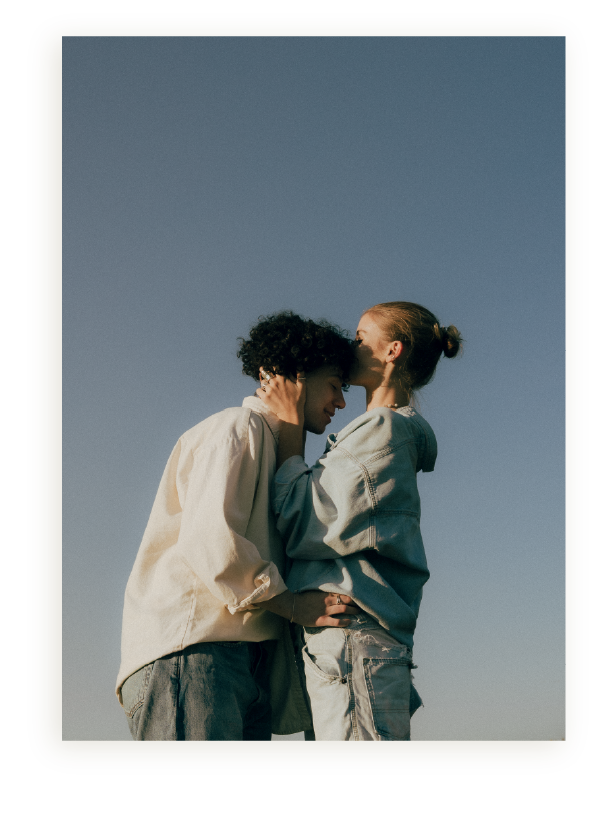 Two women hugging outdoors against a clear blue sky, one kissing the other's forehead.