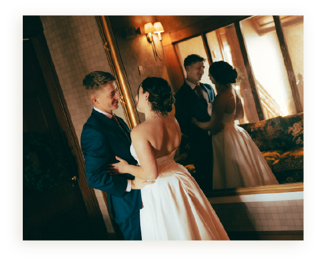 A couple dancing closely in a mirror, with another couple dancing in the background, in a warmly lit room.