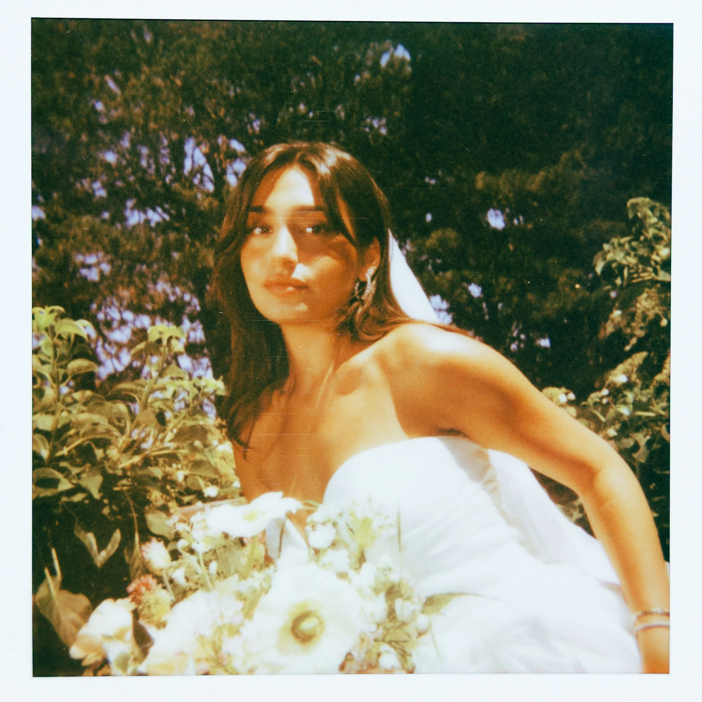 A woman with long dark hair taking a selfie outdoors in front of trees and foliage, wearing a white top.