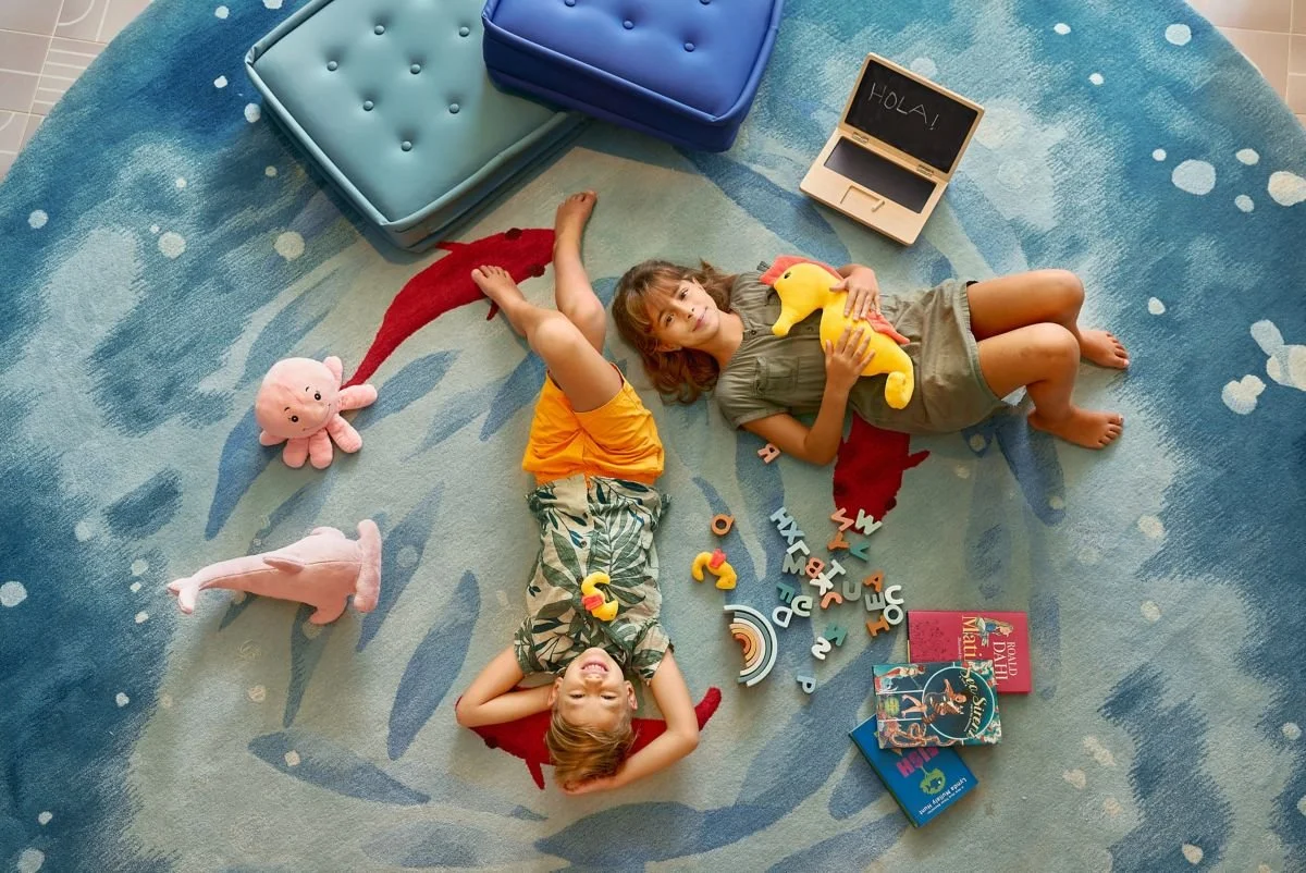 Two children lying on a blue ocean themed rug, surrounded by toys, books, and a small chalkboard with 'Hola!' written on it.
