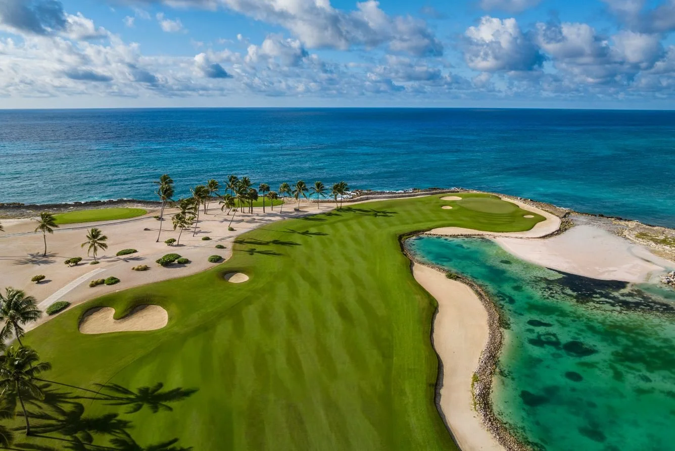 Aerial view of a seaside golf course with lush green fairways, sandy bunkers, a small pond, and rows of palm trees, with the ocean and a partly cloudy sky in the background.