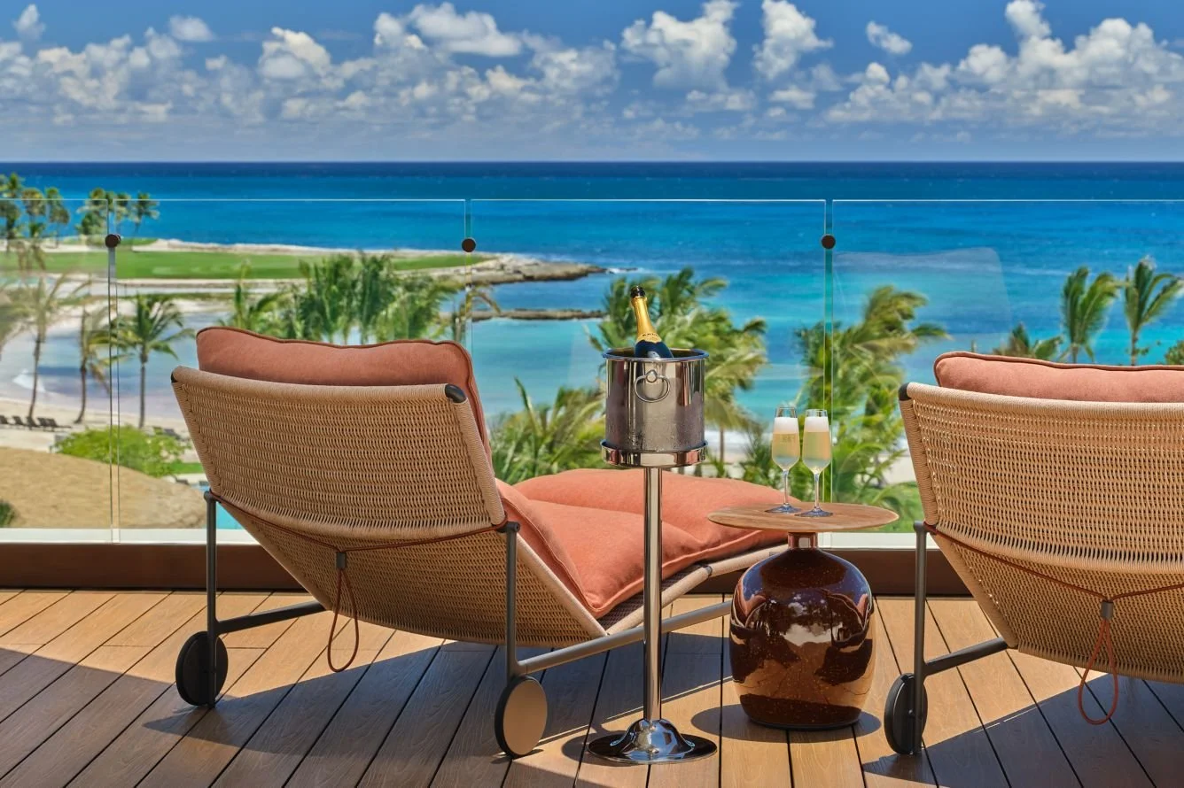 A balcony with two lounge chairs overlooking a tropical beach with palm trees, turquoise water, and blue sky with clouds. A small table holds a champagne bucket with a bottle and two glasses.