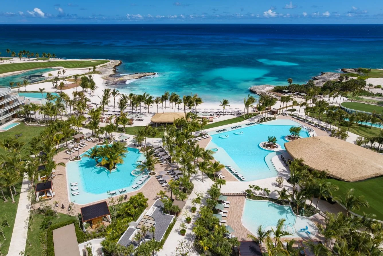 Aerial view of a tropical resort with multiple swimming pools, surrounded by palm trees, with a beach and ocean in the background.