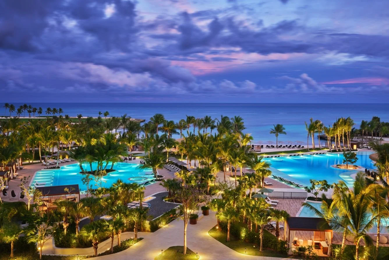 Aerial view of a tropical resort at dusk with illuminated swimming pools, numerous palm trees, lounge chairs, and a view of the ocean in the background under a cloudy sky.