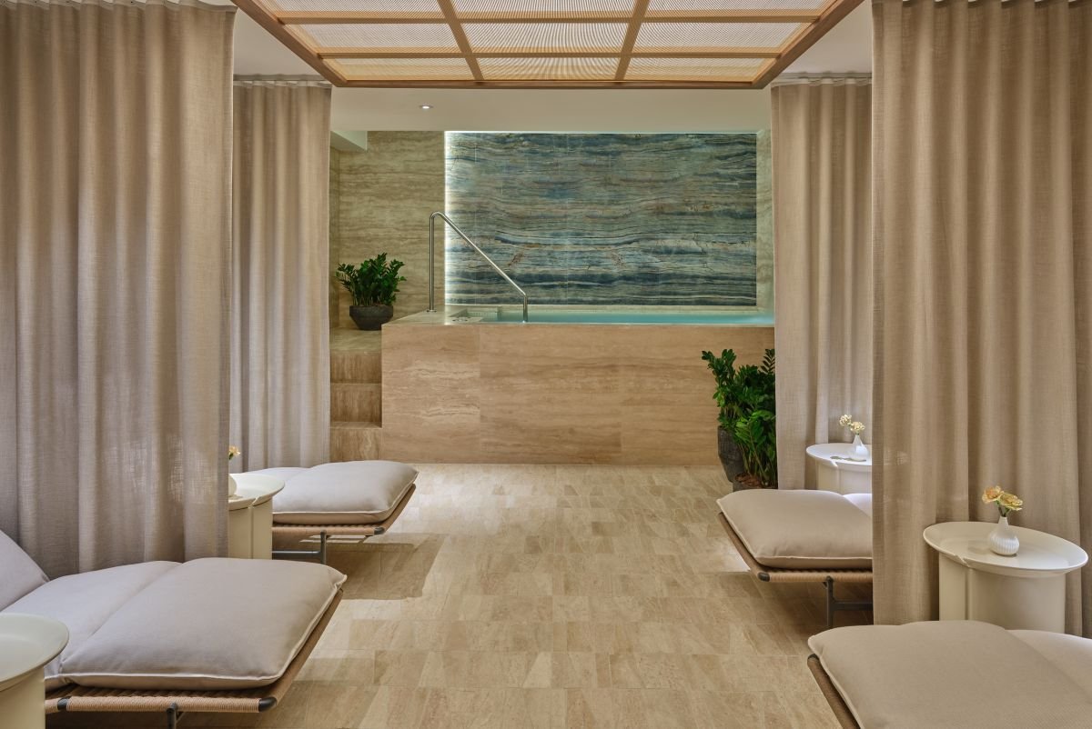 Spa room with curtained partitions, lounge chairs, small side tables with vases, potted plants, and a hot tub with a stone wall background.