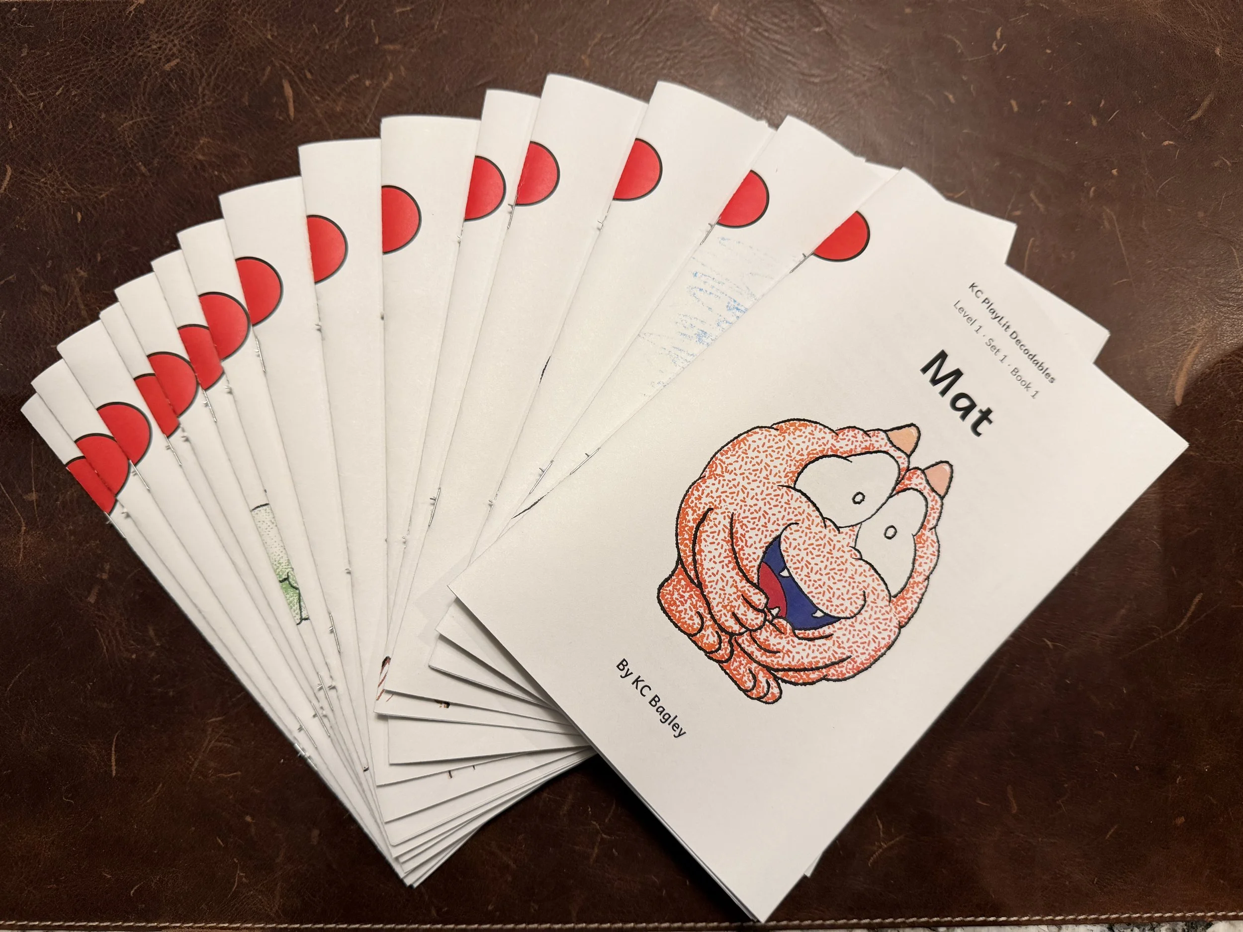 "KC PlayLit Decodable Set 1 all 15 books fanned out on leather background".  KC Bagley.  KCPlayLit. Science of reading. Bridging Play and Literacy.  UFLI aligned.