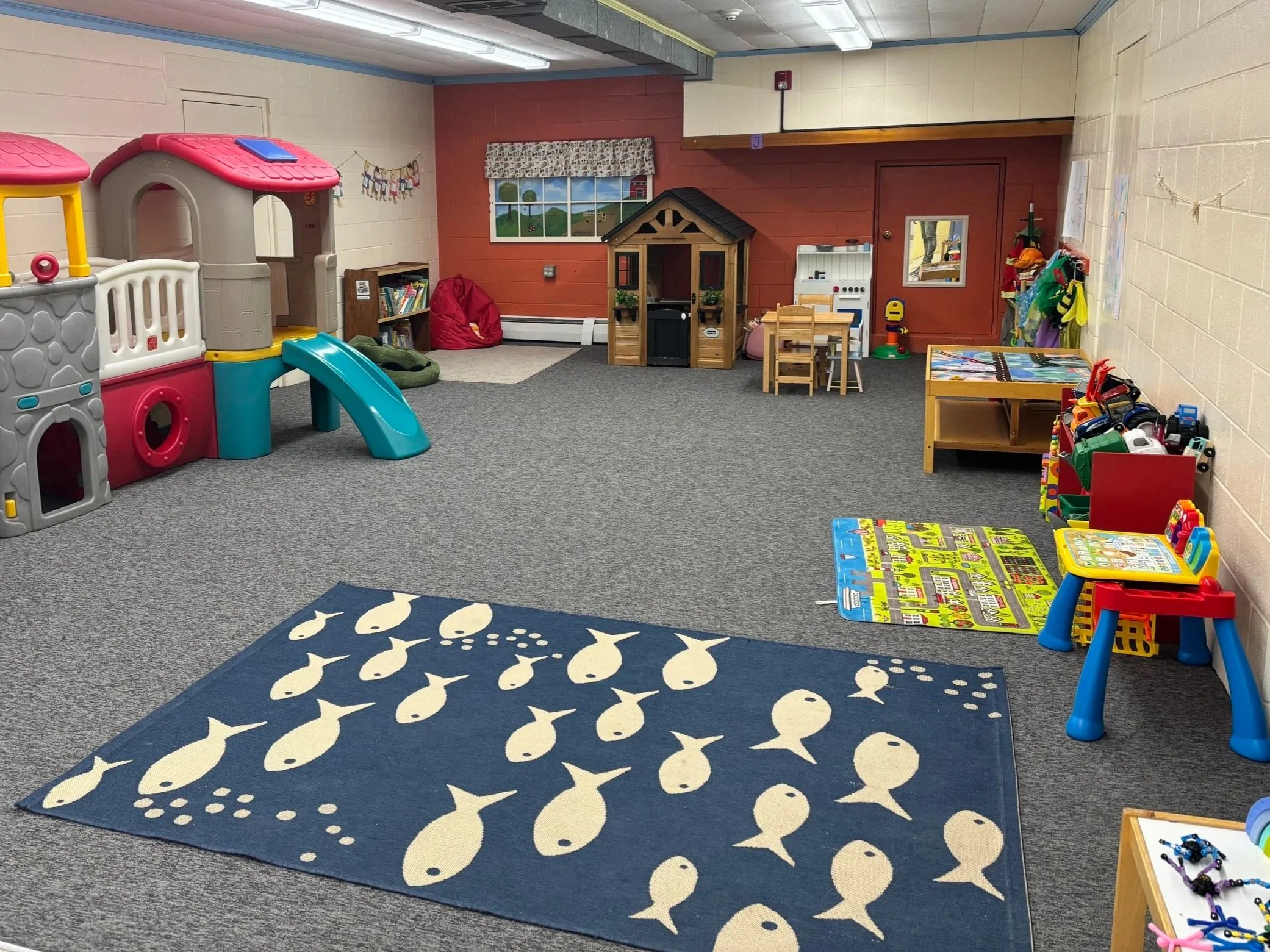 Empty indoor children's play area with a blue area rug with fish pattern in the foreground, plastic playhouse with slide on the left, play kitchen, small tables and chairs, bookshelves, and various toys and decorations along the walls.