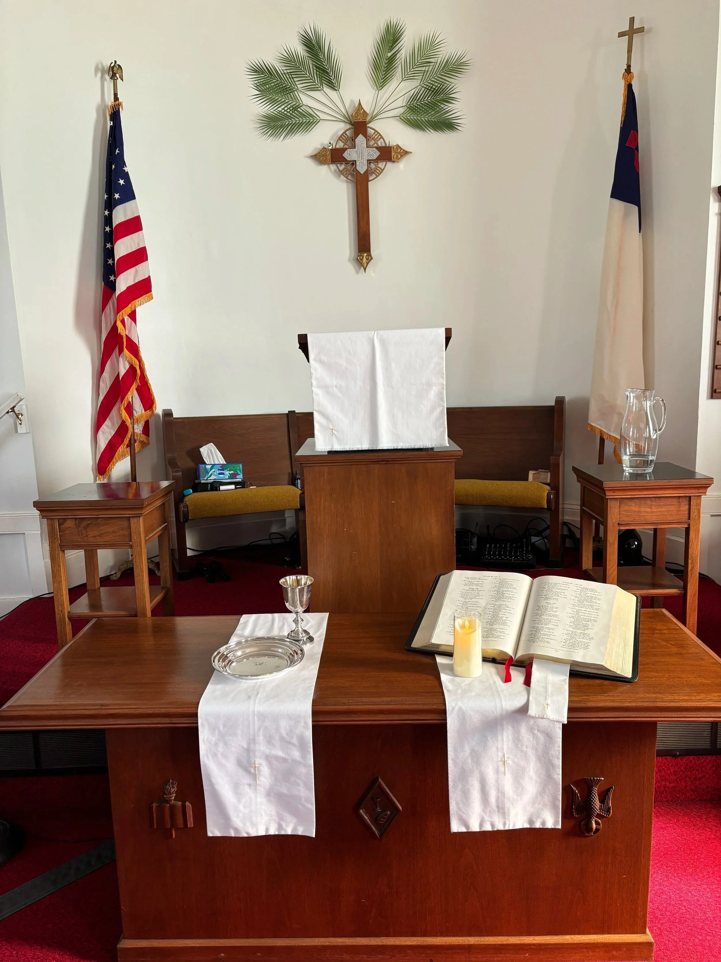 Interior of a Christian church altar with flags, a cross, an open Bible, candles, a chalice, and religious symbols.