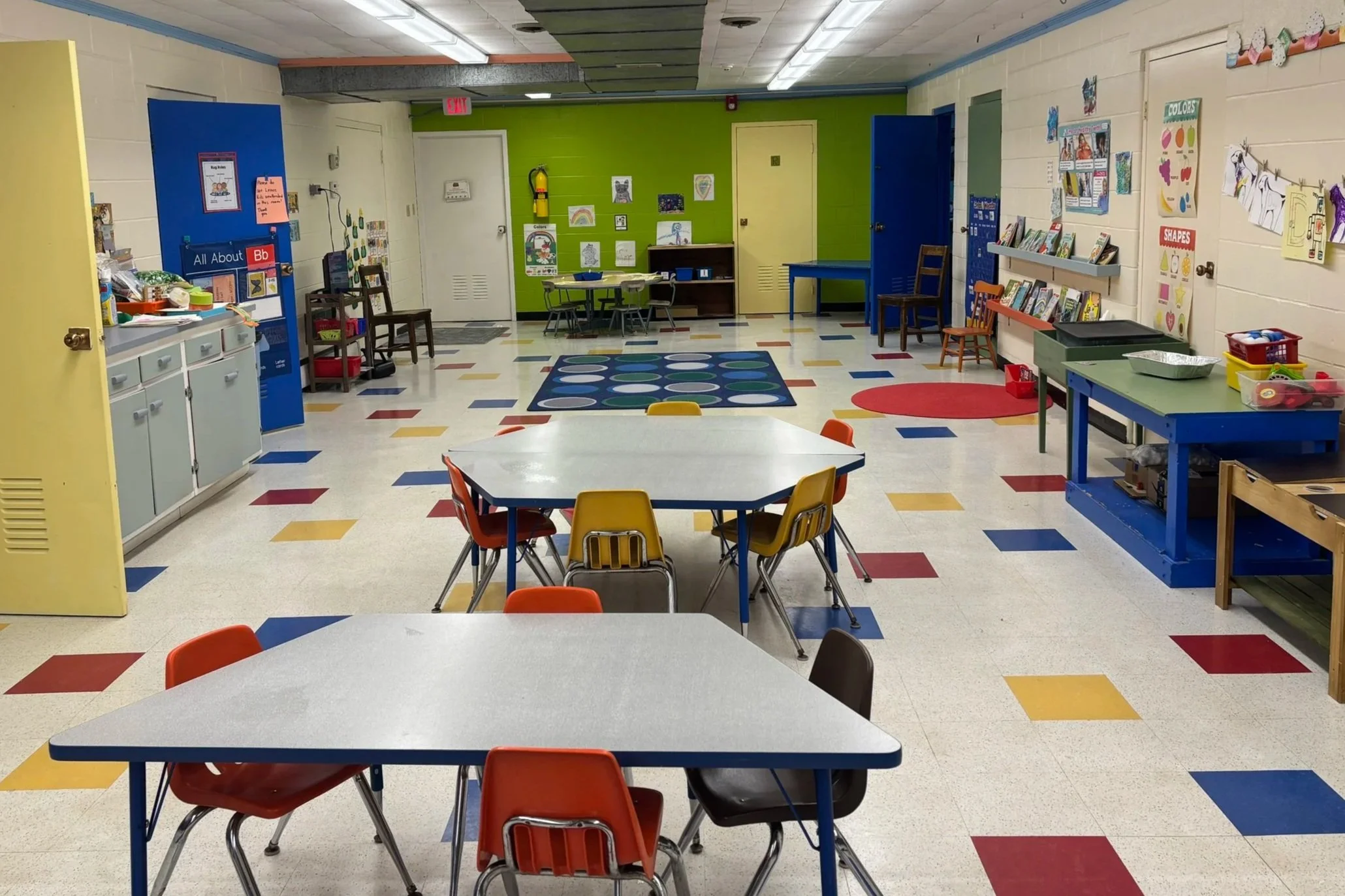 Colorful preschool classroom with child-sized tables and chairs, educational posters, and children's artwork on the walls.