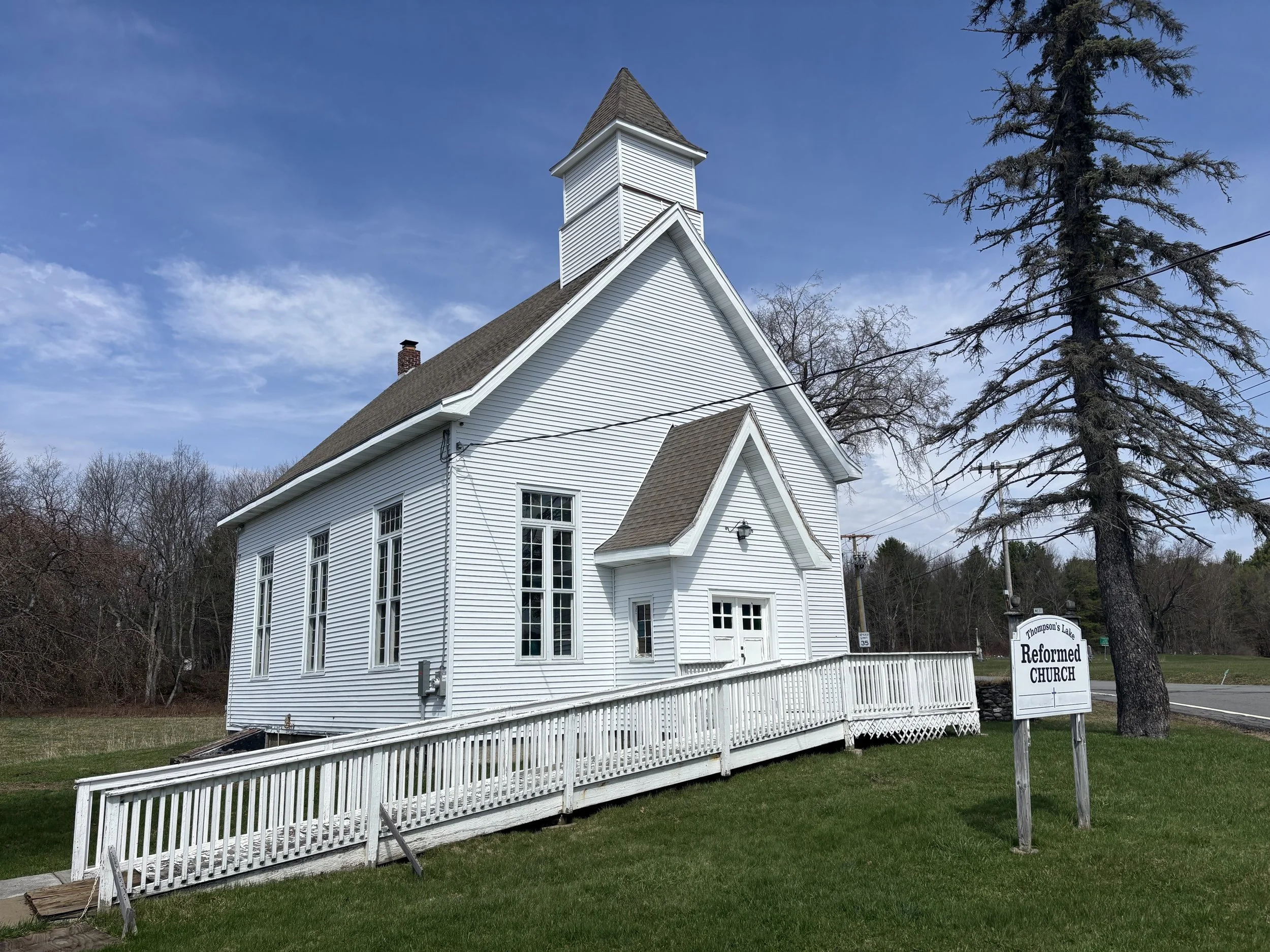 A white church with a small steeple, brown shingled roof, and a white wooden ramp leading to the entrance, surrounded by a grassy lawn.