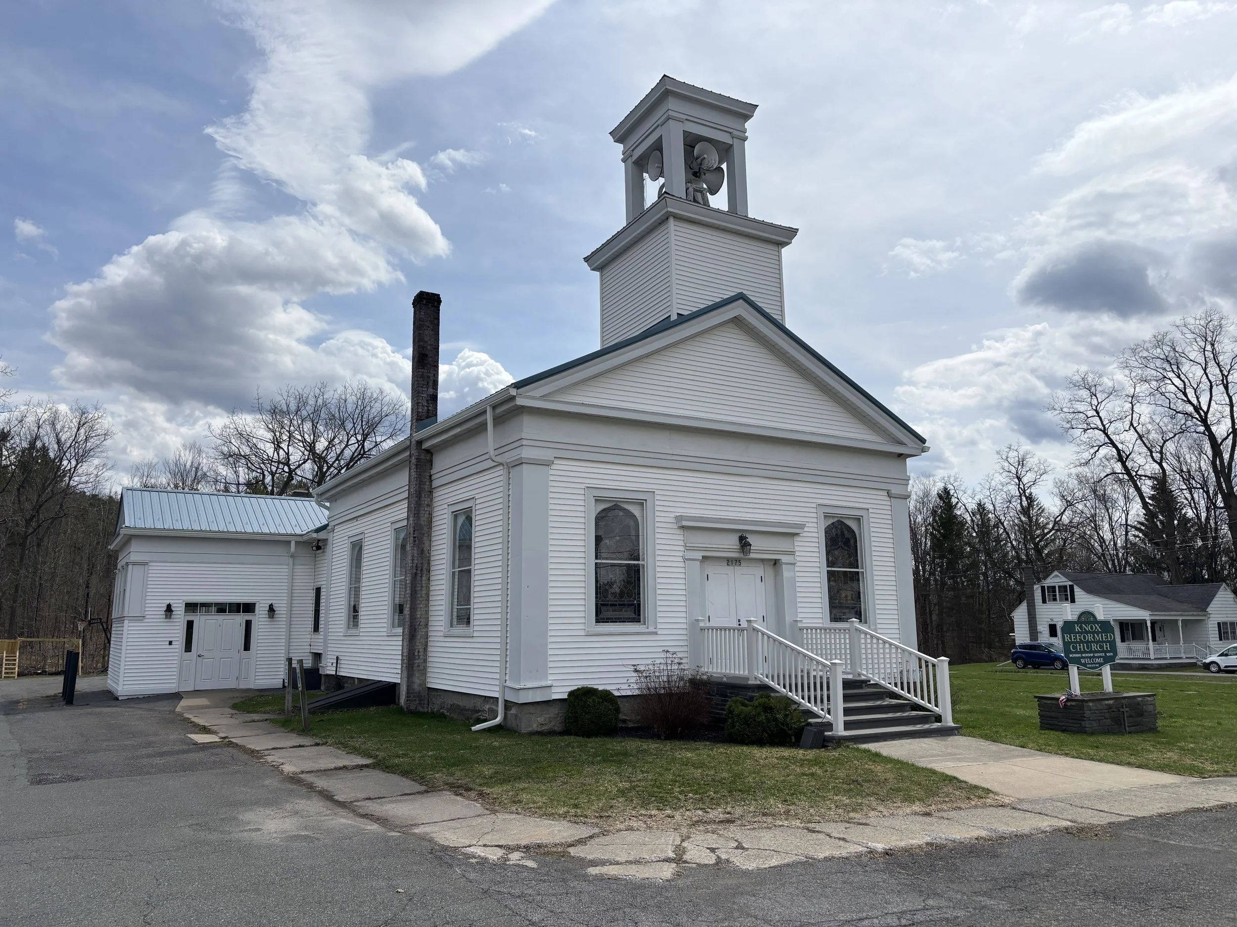 White church with steeple, front stairs, and a sign that reads 'Knox Reformed Church' in front, surrounded by grass and trees, under partly cloudy sky.
