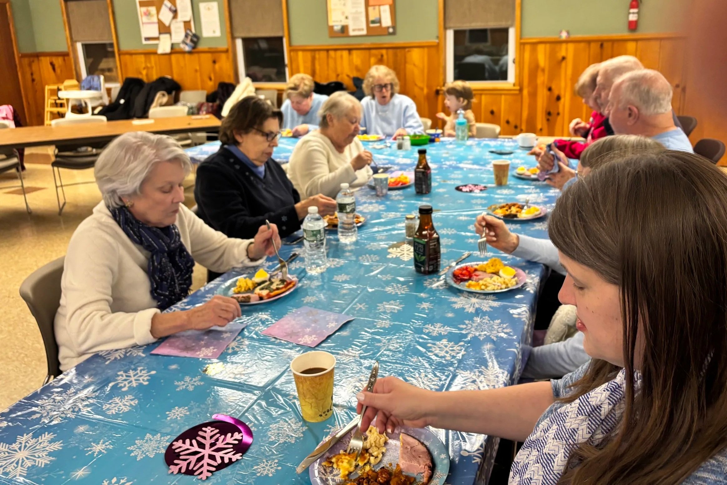 Group of elderly and middle-aged people sitting at a long table, enjoying a meal in a room decorated with holiday-themed tablecloths and pink snowflake decorations.