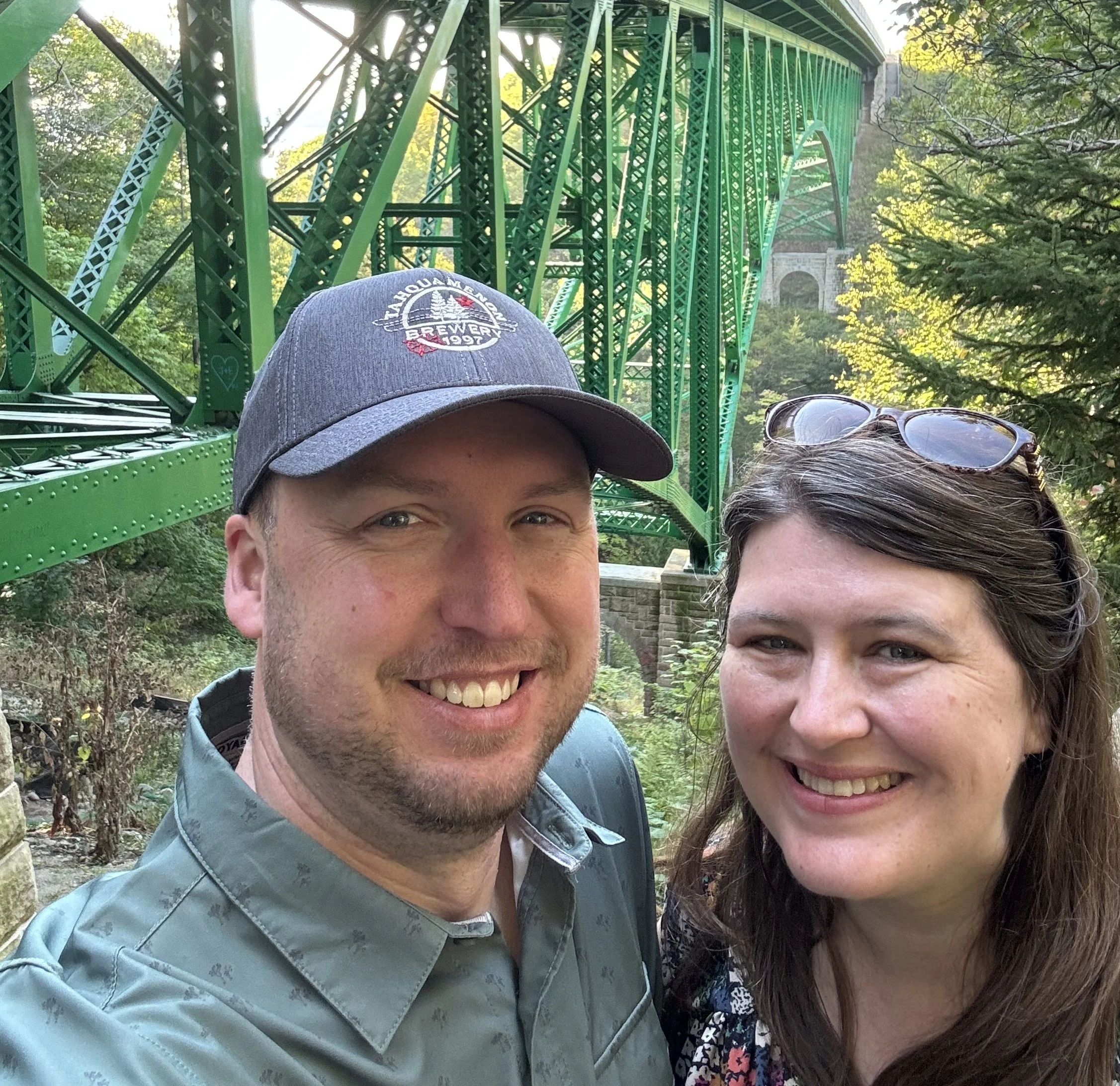 A smiling man and woman taking a selfie outdoors near a green metal bridge. The man wears a gray baseball cap and a gray shirt, and the woman has brown hair and sunglasses on her head. There are trees and greenery around them.