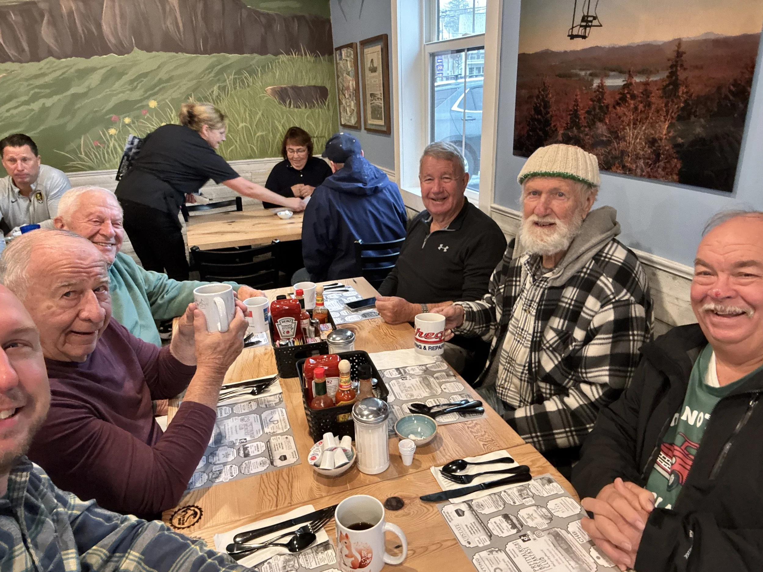 A group of elderly men and women sitting at a restaurant table, smiling, with condiments and drinks on the table, and a mural of nature on the wall in the background.