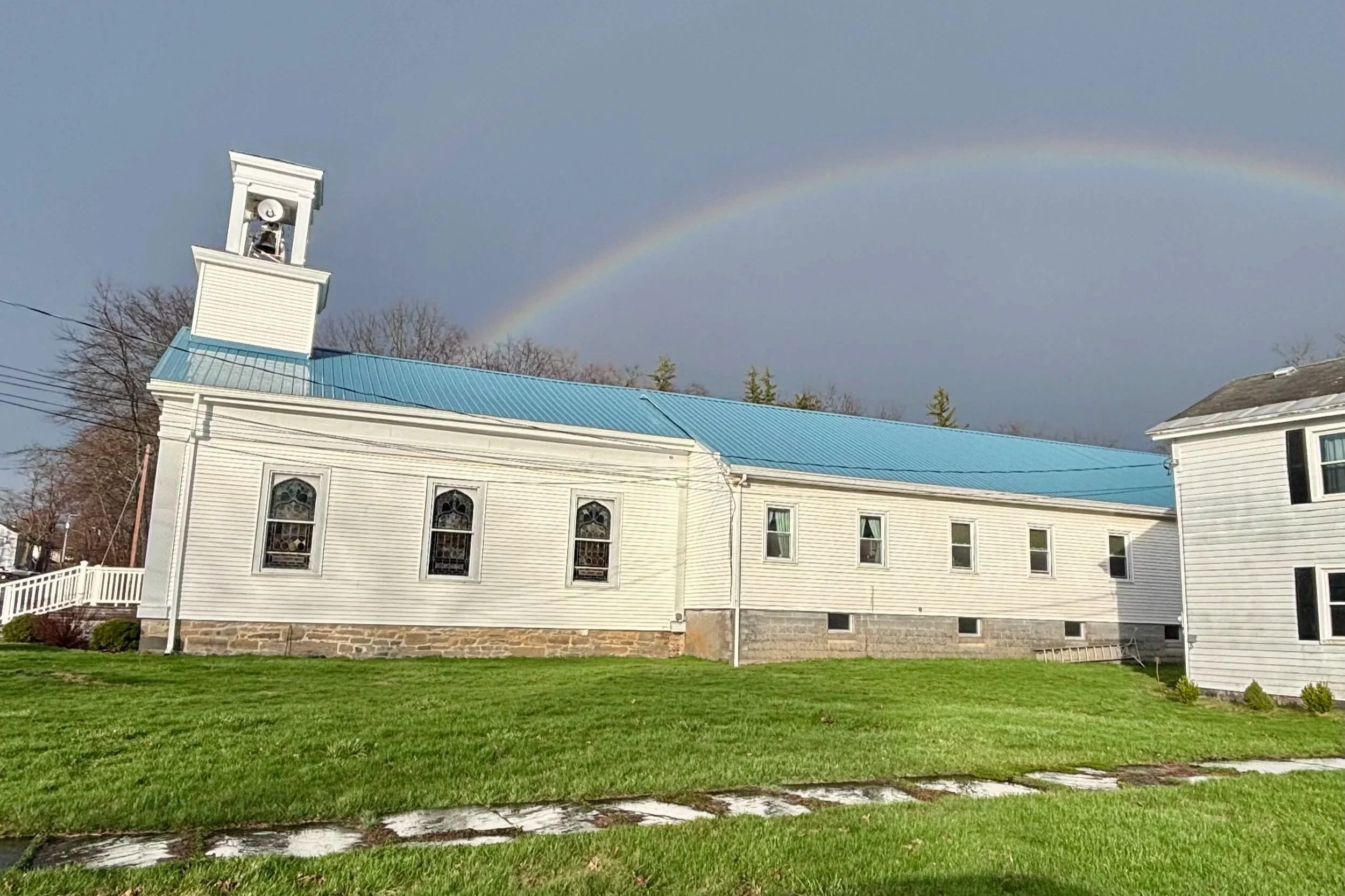 White church building with a blue roof and stained glass windows, a small bell tower on the left, green grass in front, and a rainbow in the sky behind.