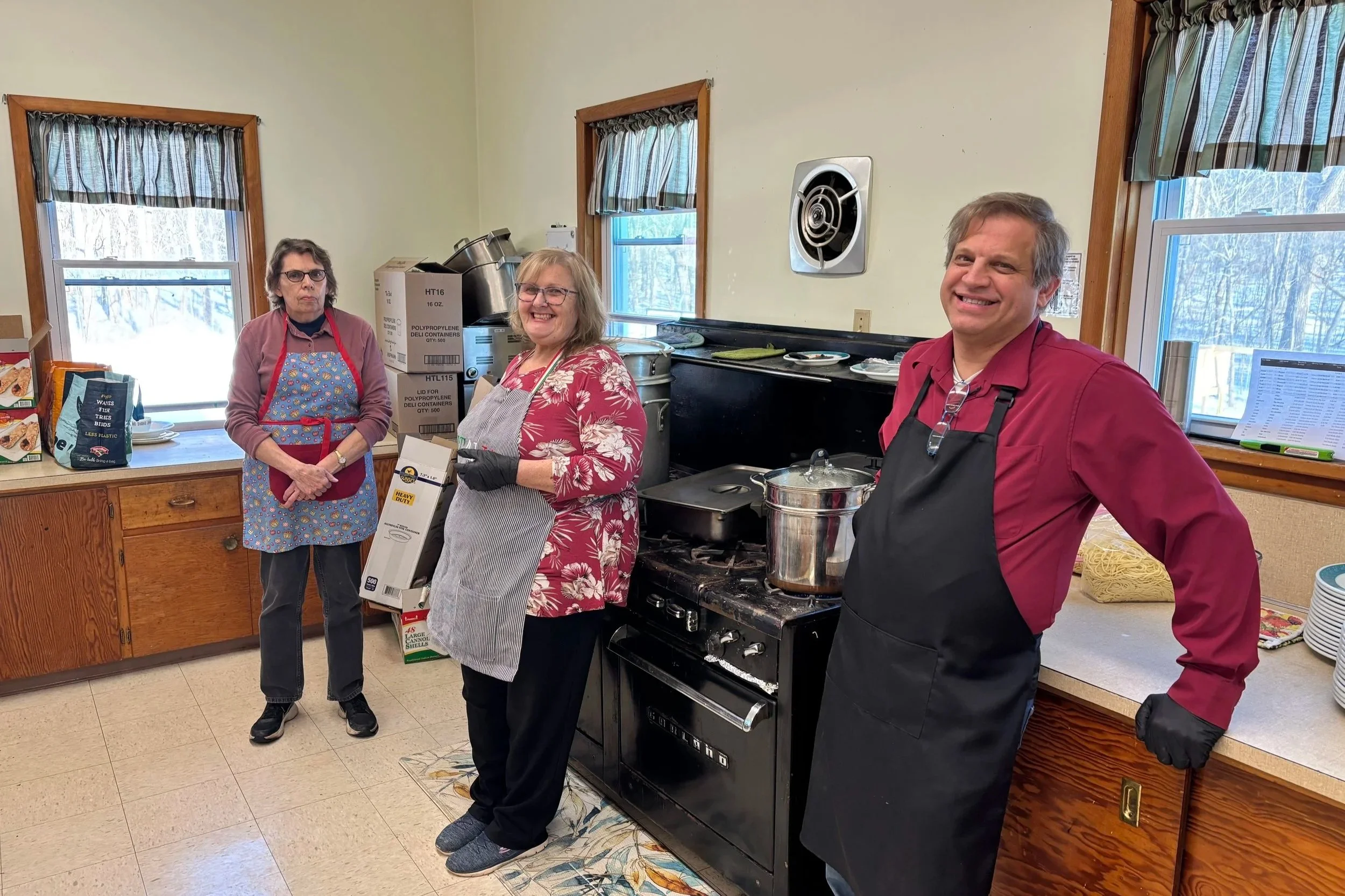 Three people in a kitchen, two women and one man, preparing food and smiling. The kitchen has two windows with striped curtains and a black stove with a large pot, surrounded by boxes and food items on the counters.