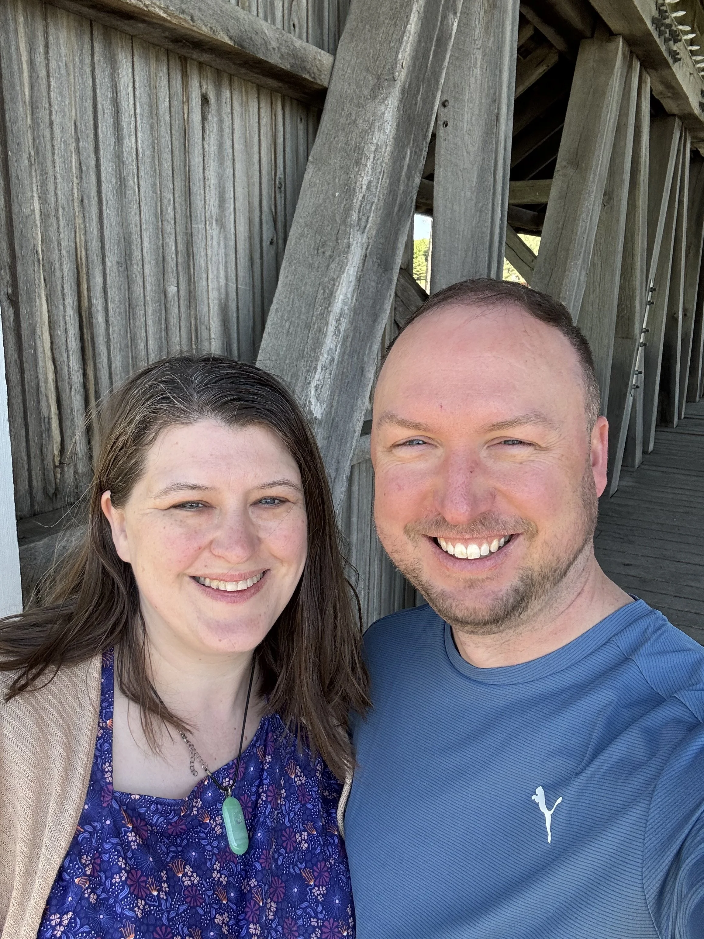 A smiling couple taking a selfie outdoors near a wooden structure on a sunny day.