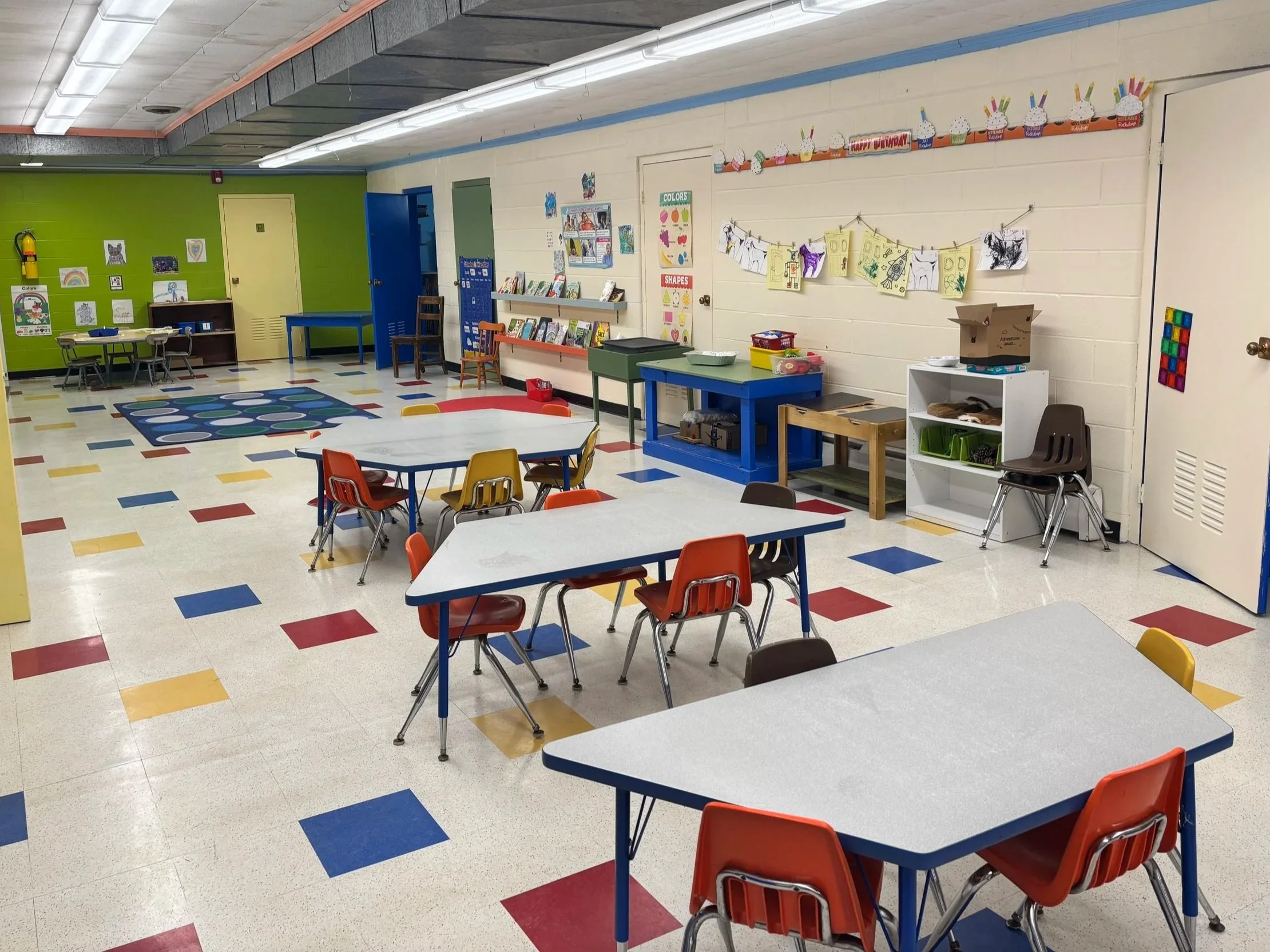 Empty classroom with colorful tables and chairs, children's artwork on the wall, and educational decorations.