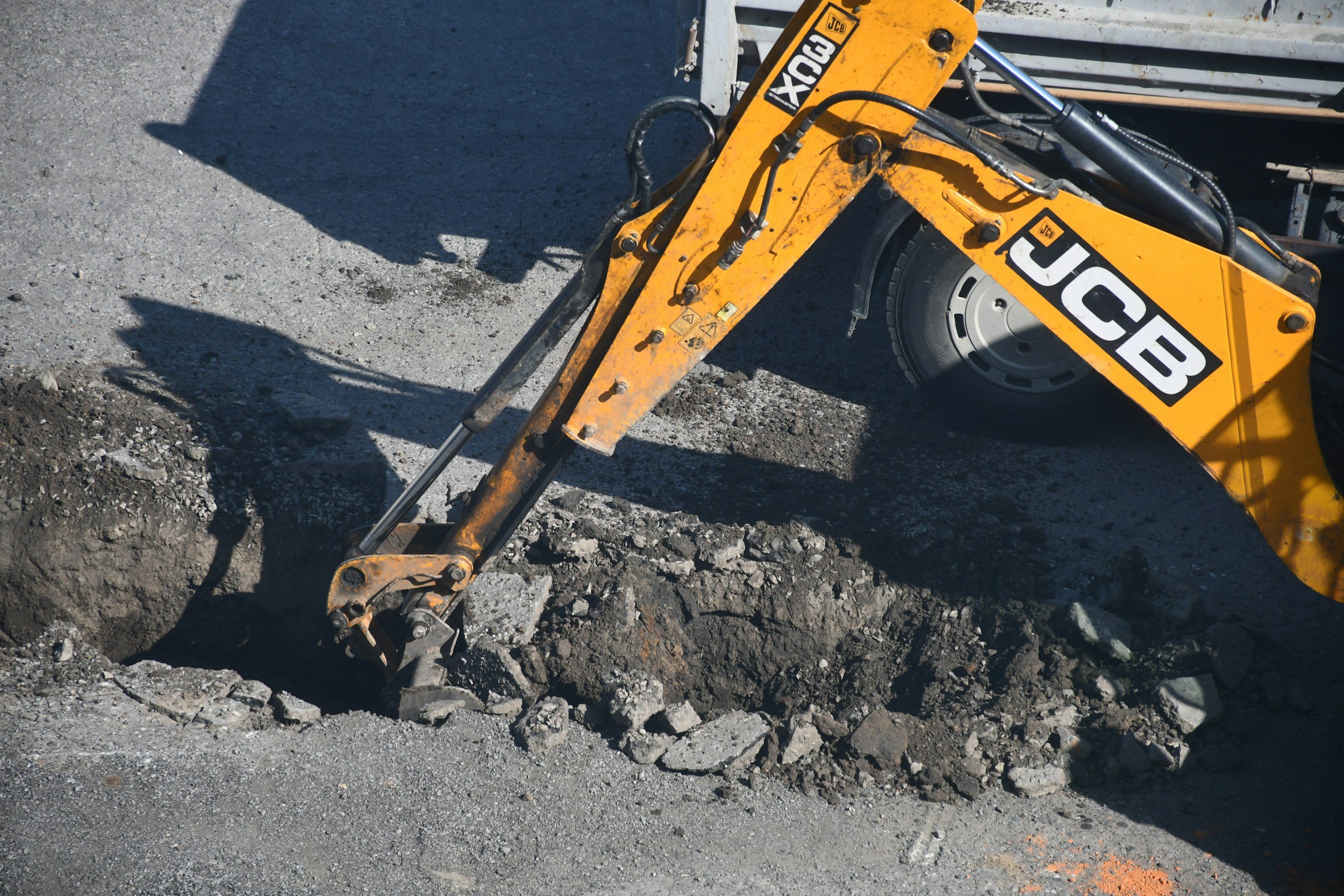 Close-up of a yellow JCB mini excavator digging into the asphalt road surface, with asphalt and dirt pile visible.