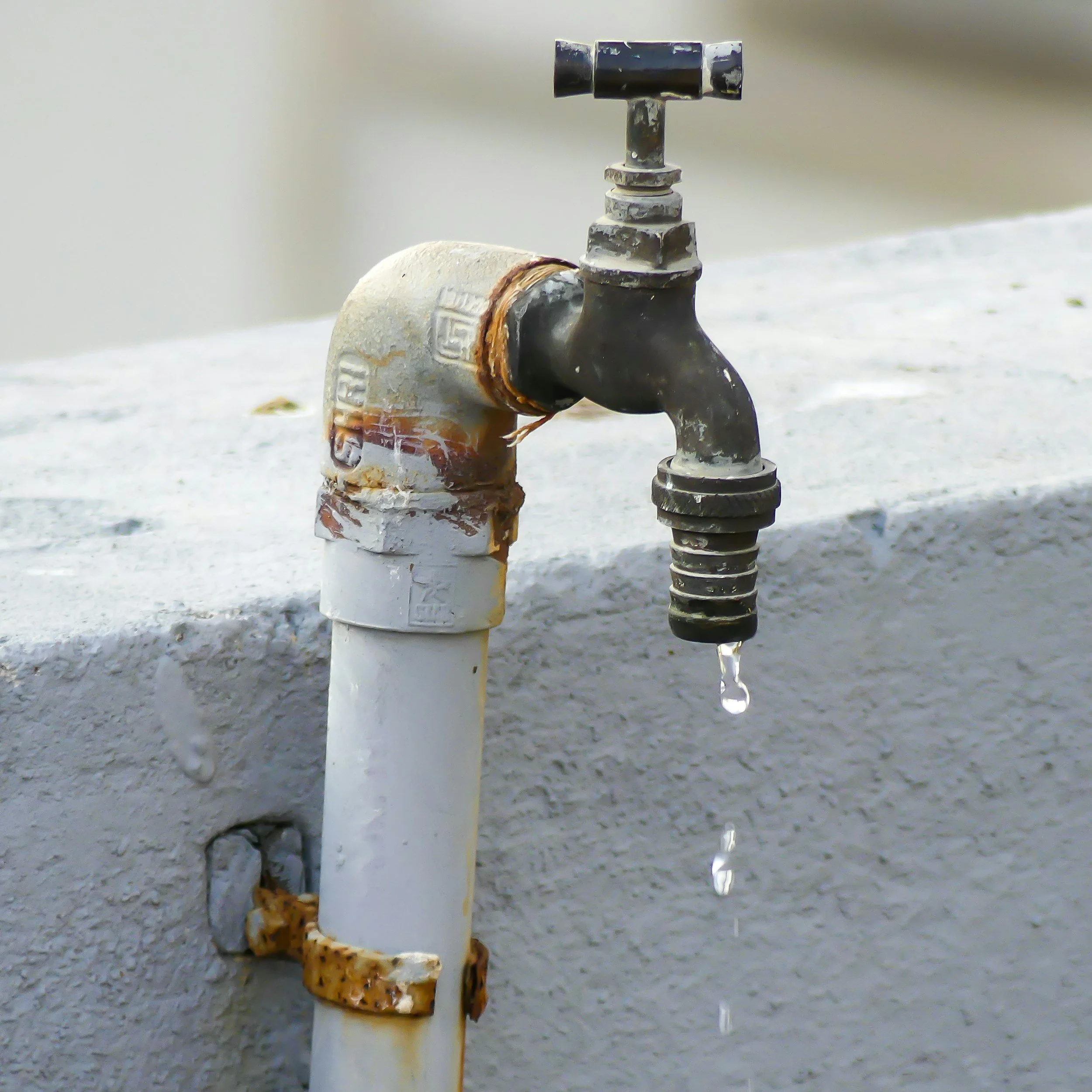 A rusty water tap dripping water over a white concrete surface.