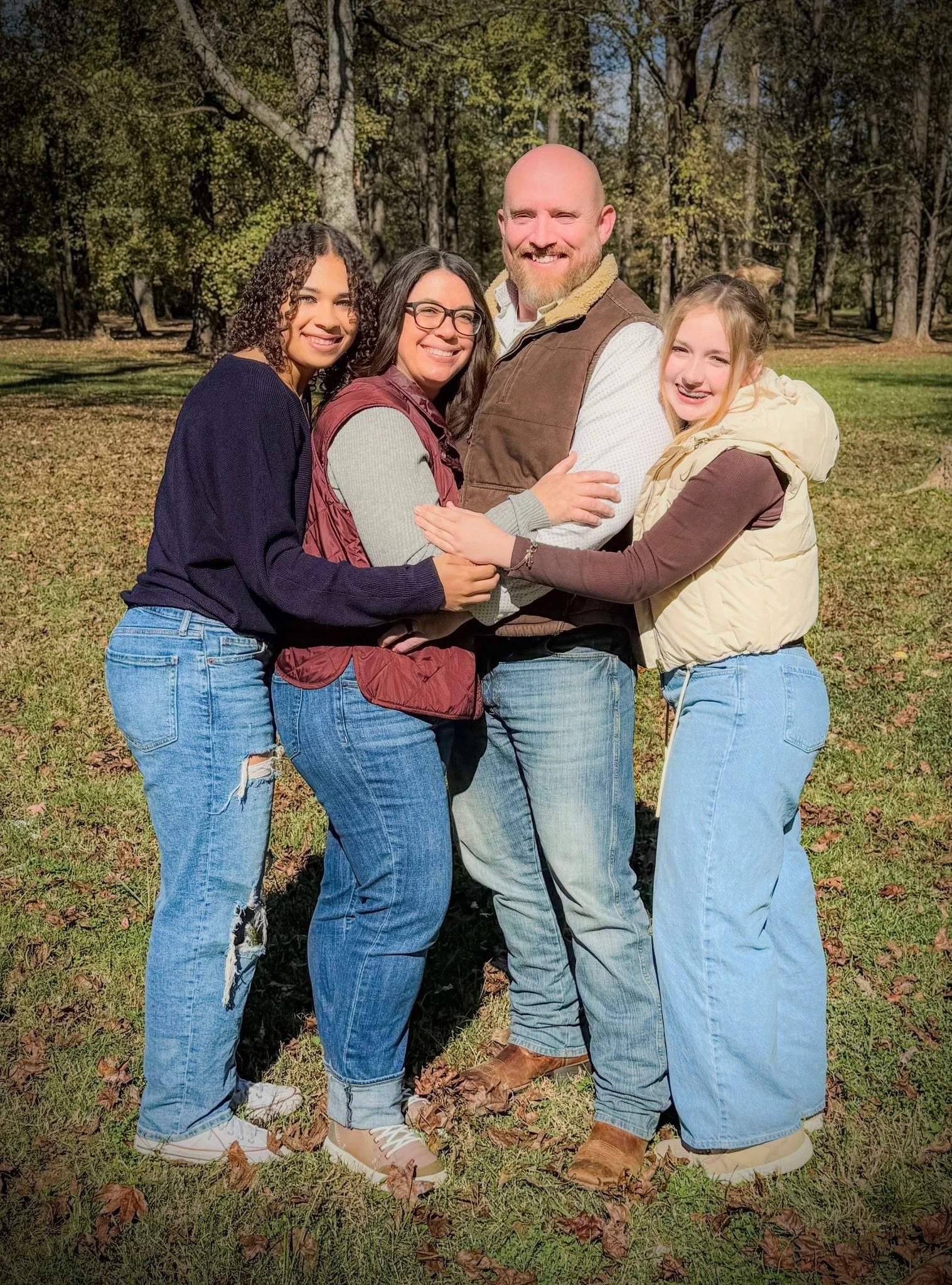 Group of four people hugging outdoors in a wooded park during daytime.