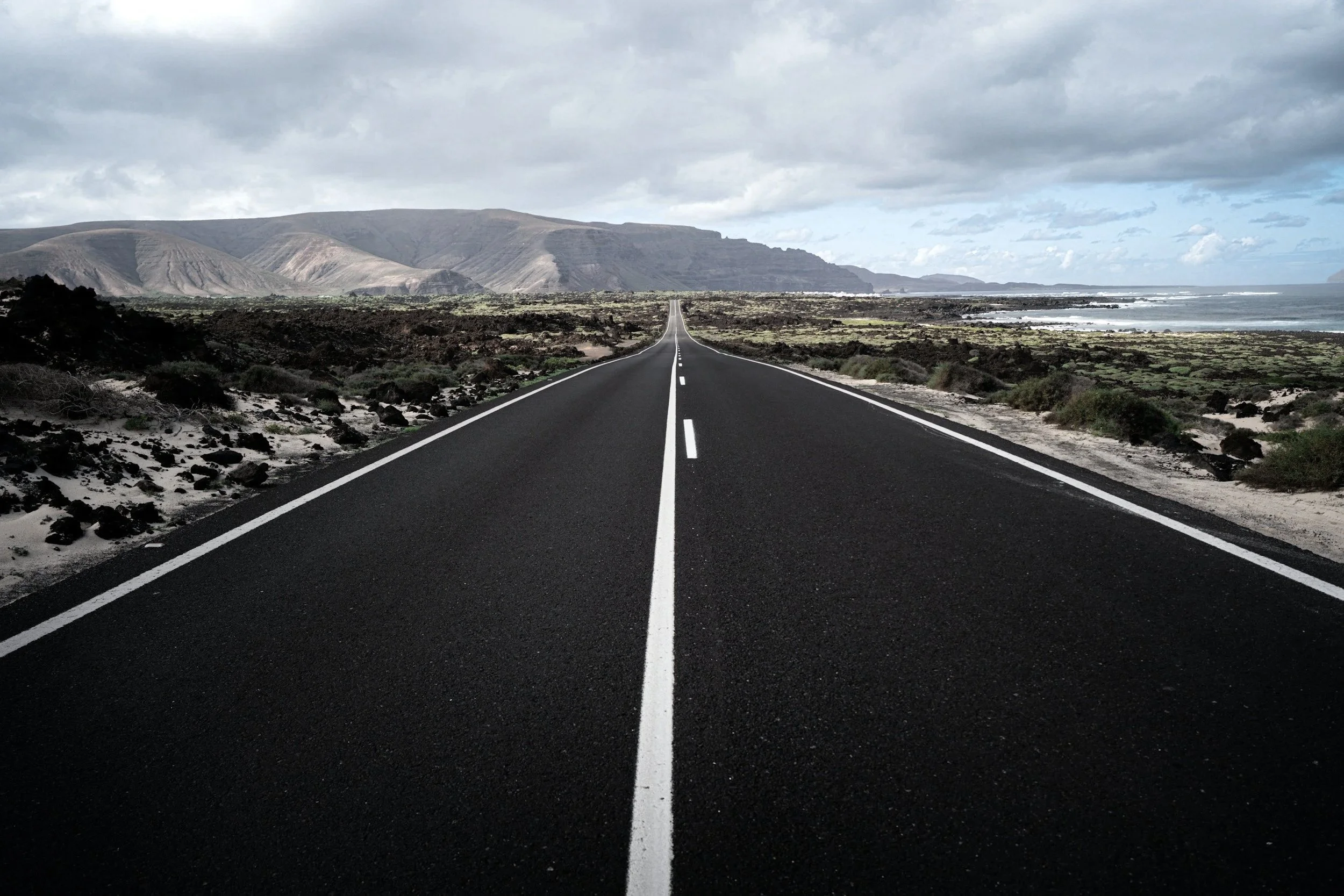 A straight, black asphalt road with white dashed center lines and solid edges, stretching through a coastal landscape with sparse vegetation, black volcanic rocks, and distant mountains under cloudy skies.