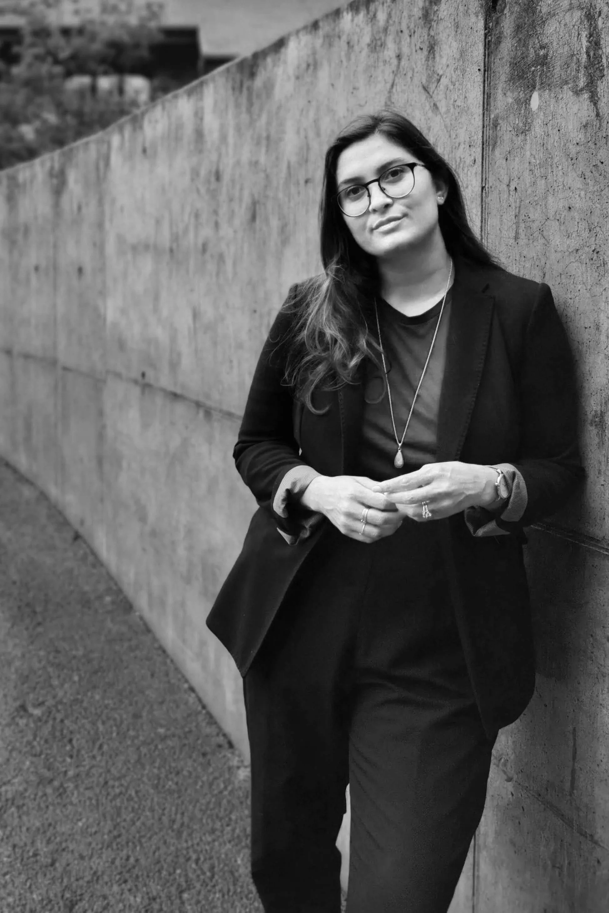 A woman with glasses standing outdoors next to a concrete wall, wearing a blazer and a necklace.