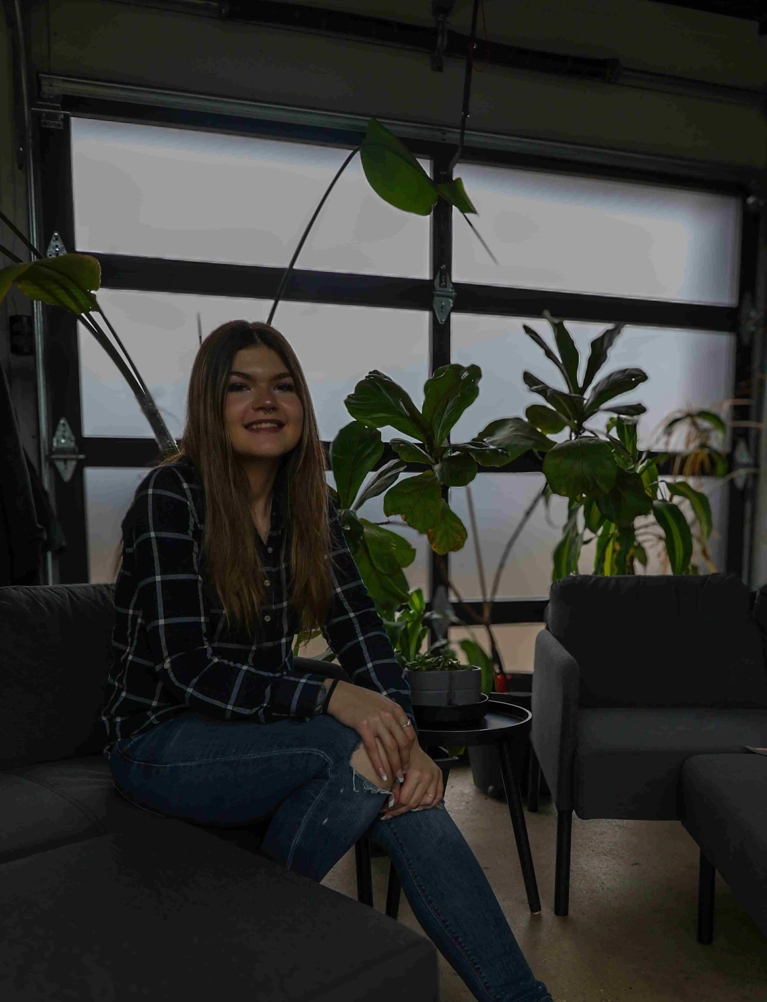 A woman sitting on a gray sofa in front of a large indoor plant with broad green leaves, inside a room with frosted glass windows.