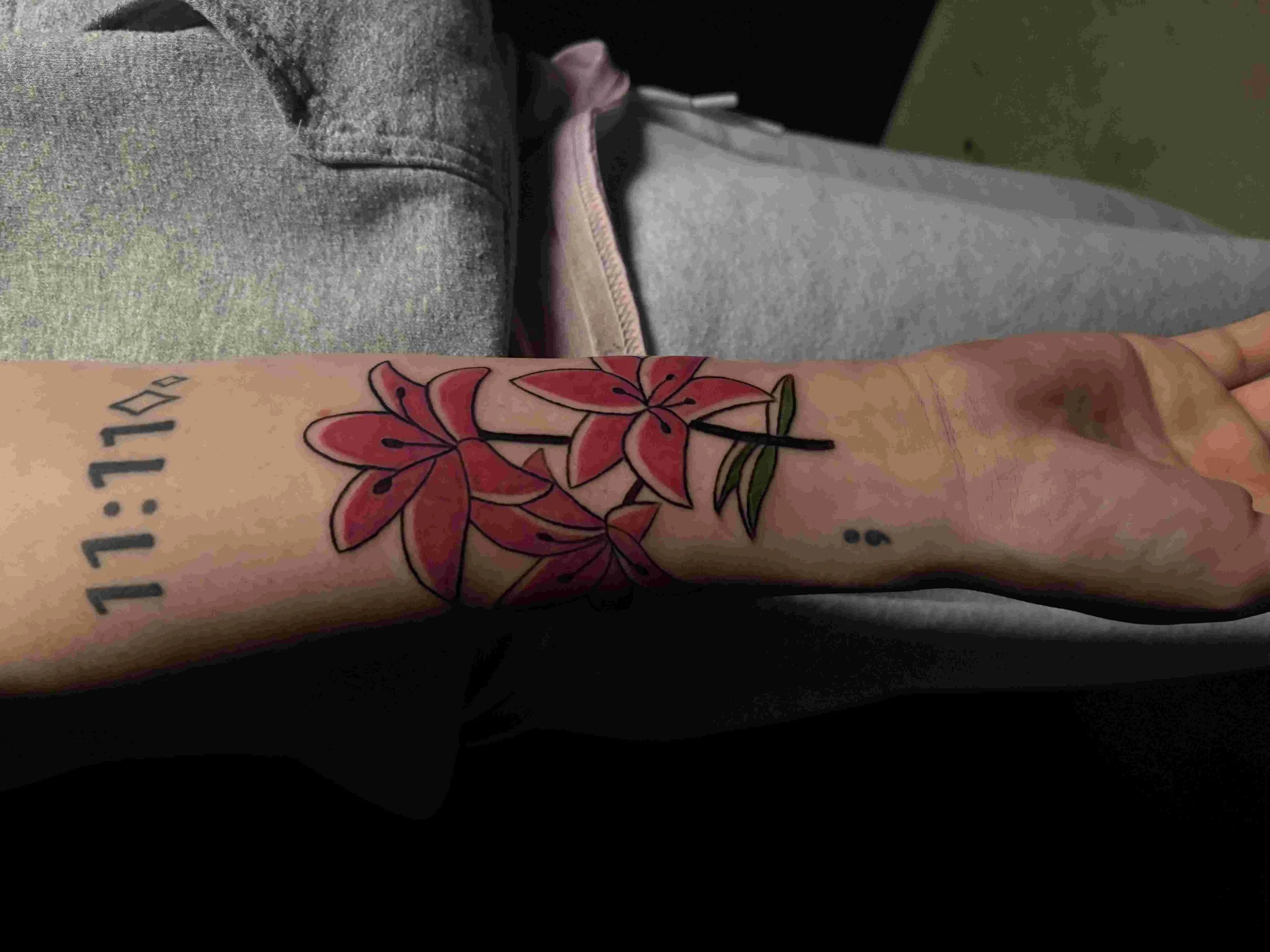 A person's arm with a colorful tattoo of red and pink leaves and green accents. The tattoo also includes the text '11:11' around the wrist.