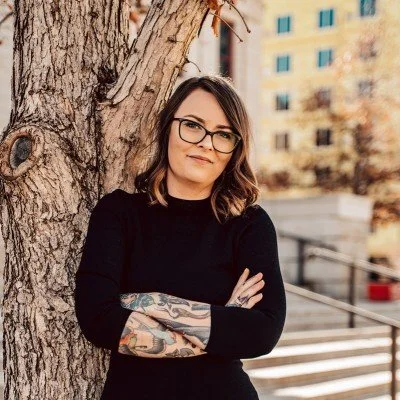 Woman with glasses and tattoos standing outdoors next to a tree, crossing her arms, with buildings in the background.