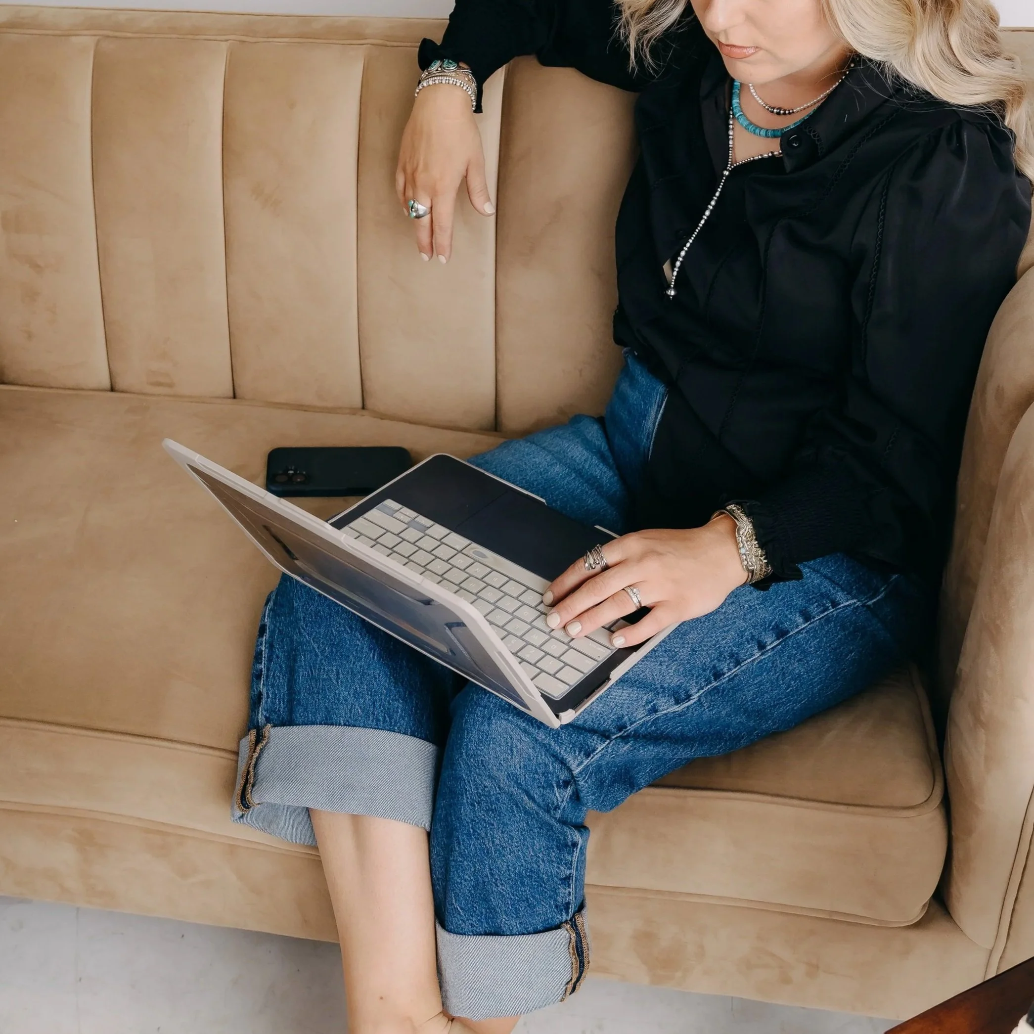 A woman sitting on a beige sofa using a small laptop, with a smartphone placed on the sofa beside her.