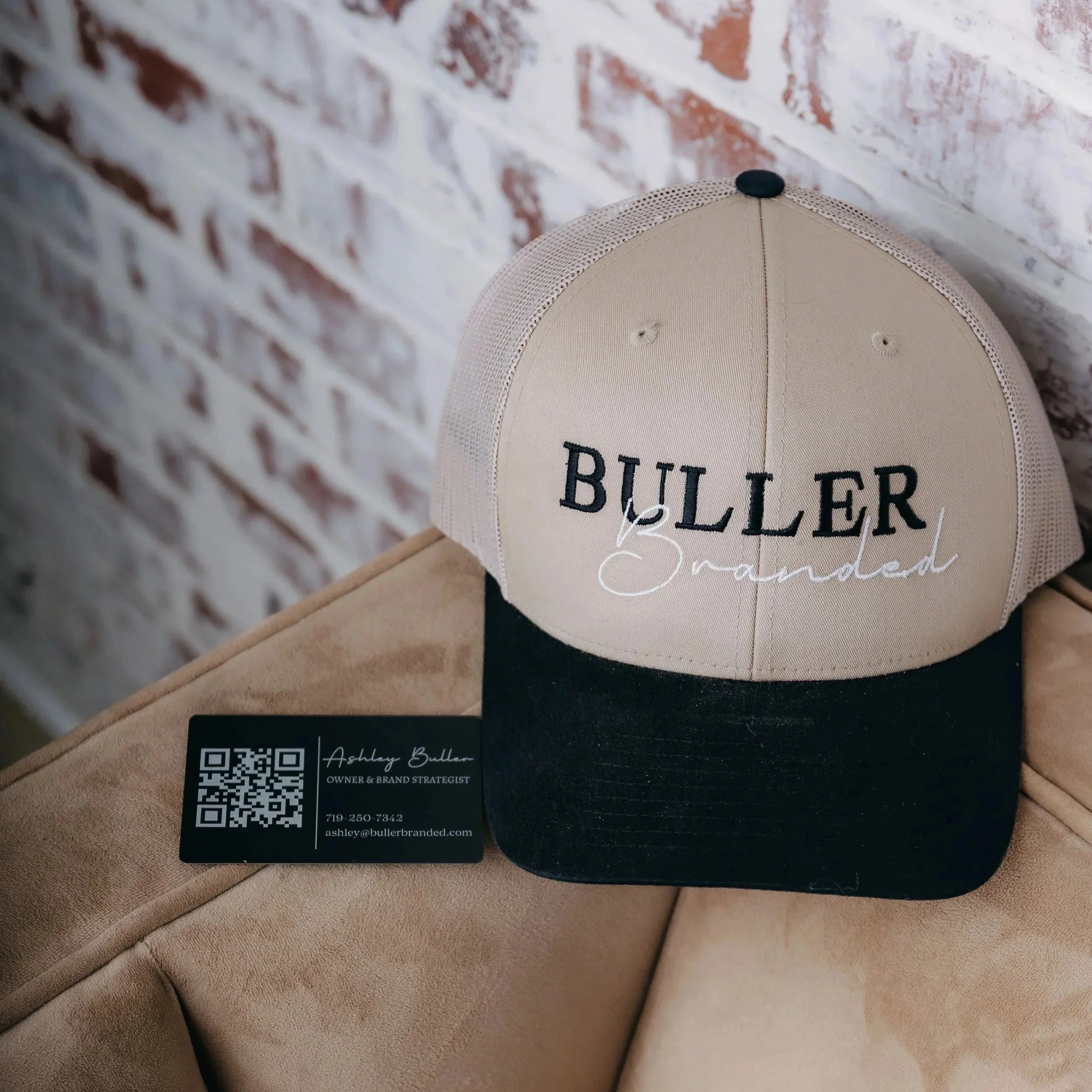 A beige and black baseball cap with the words 'BULLER' embroidered on the front and 'Branded' in cursive below. The cap is placed on a beige fabric surface, next to a business card with a QR code and the contact information for Ashley Buller, Owner & Brand Strategist.