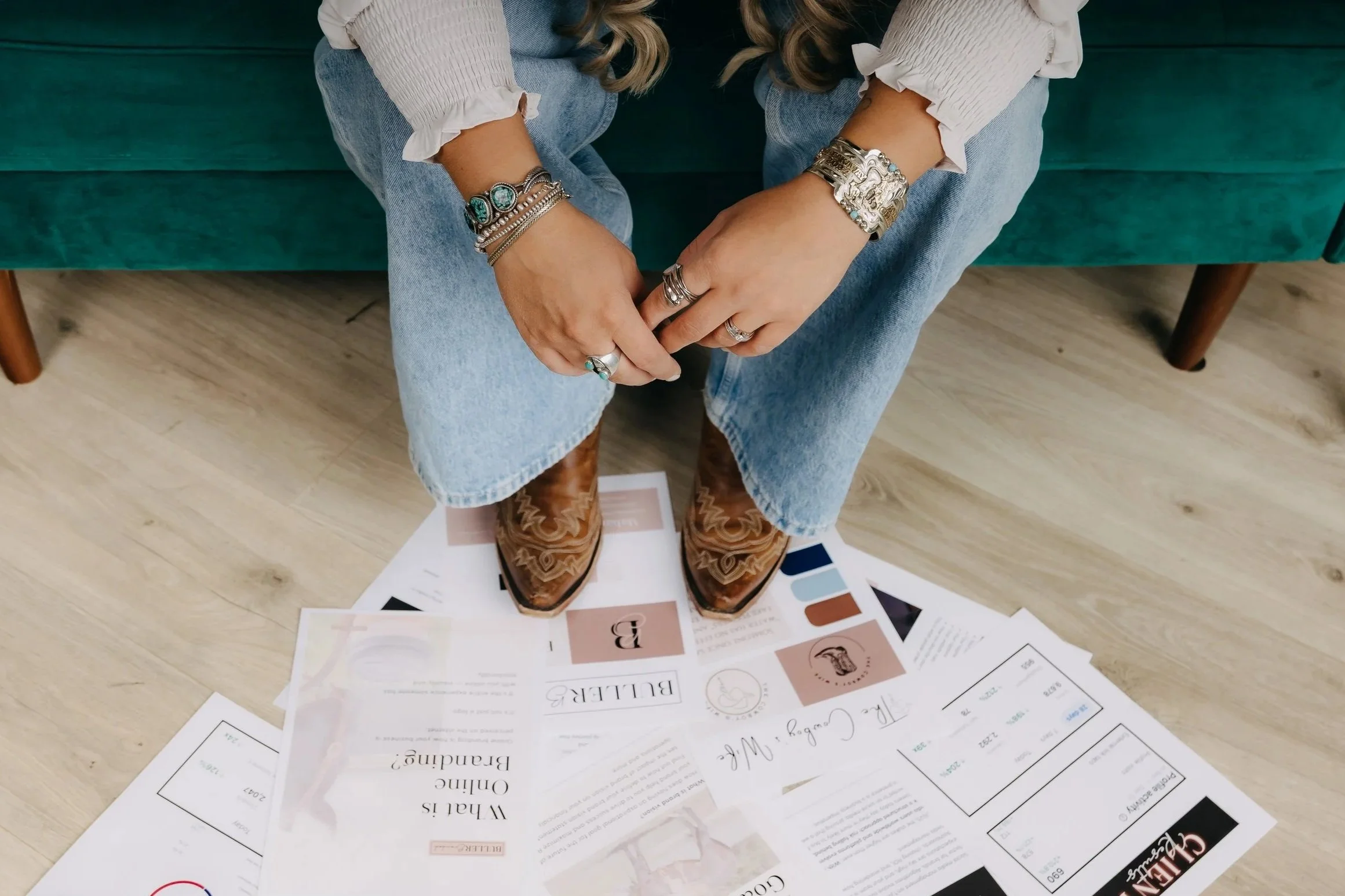 Person sitting on a green couch, wearing cowboy boots, jeans, and a white blouse, with multiple rings and bracelets on their hands, holding their hands together, with papers and brochures spread on the floor beneath their feet.