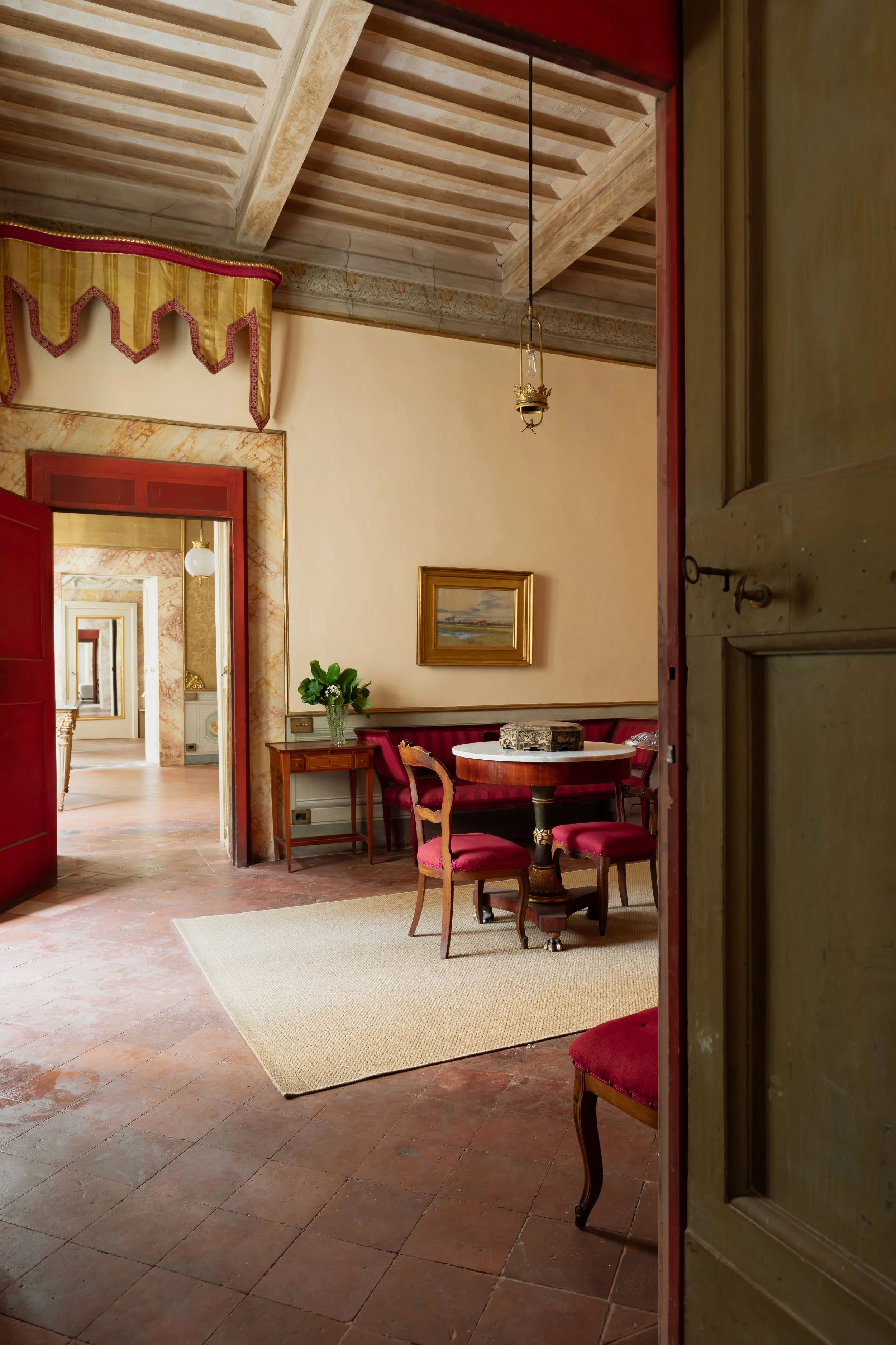 View of a vintage dining room with a round table, red cushioned chairs, a sideboard with a plant, framed painting, and patterned curtains, seen through a partially open door.