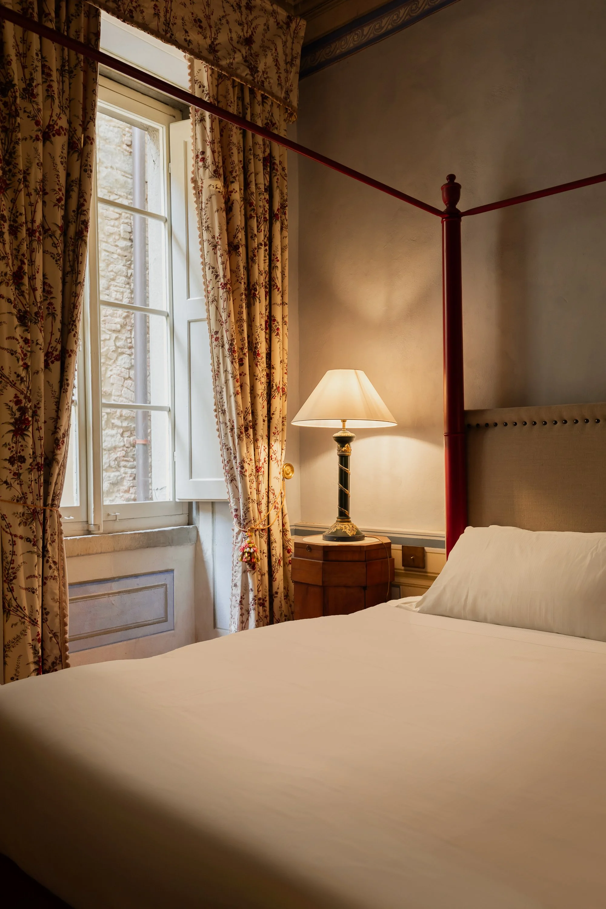 A neatly made bed with white sheets and a pillow in a cozy bedroom, next to a window with floral curtains and a lit table lamp on a wooden nightstand.