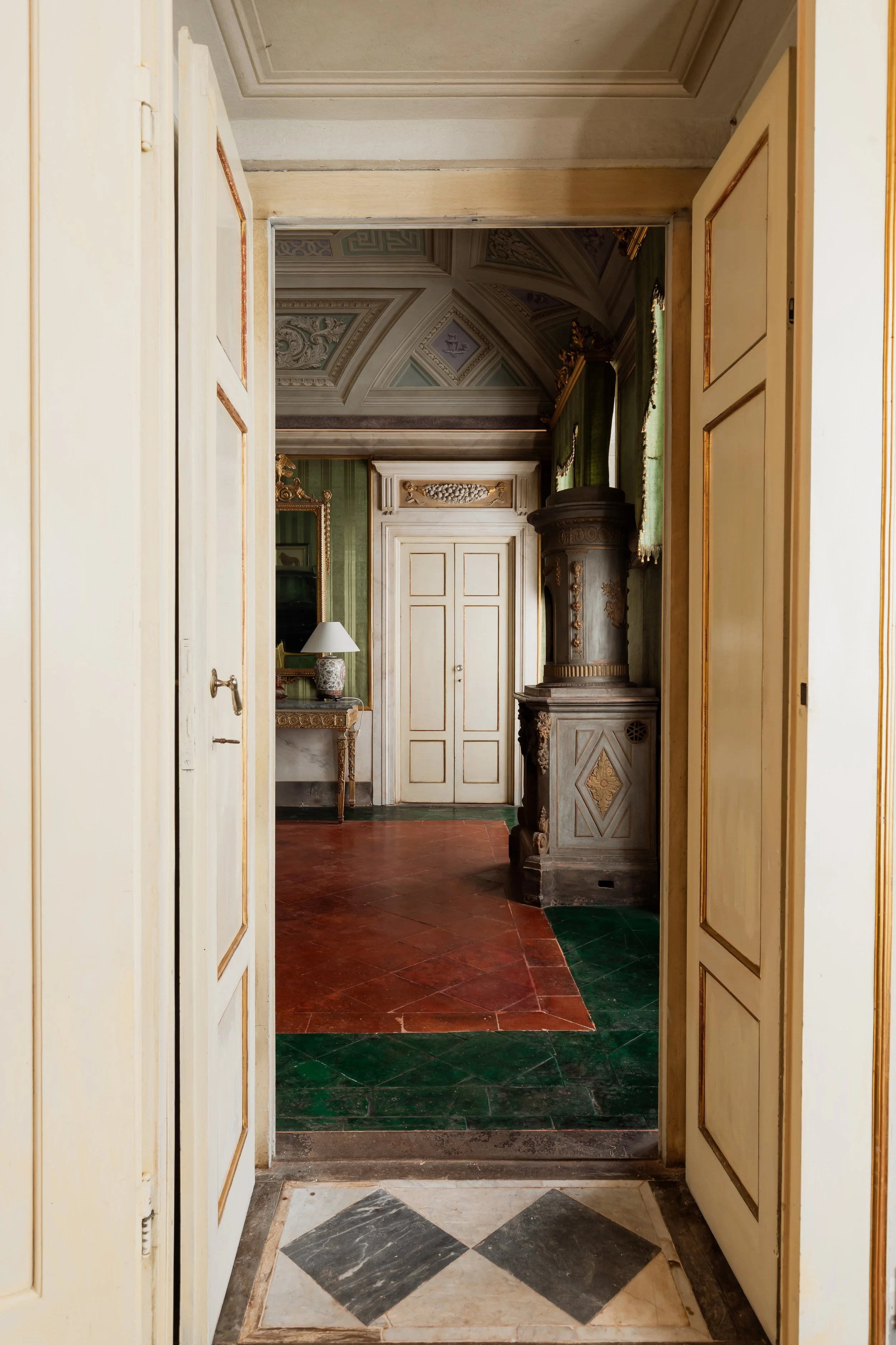 View through open double doors into an elegant room with ornate ceiling, green drapes, a vintage fireplace, and a small table with a lamp.