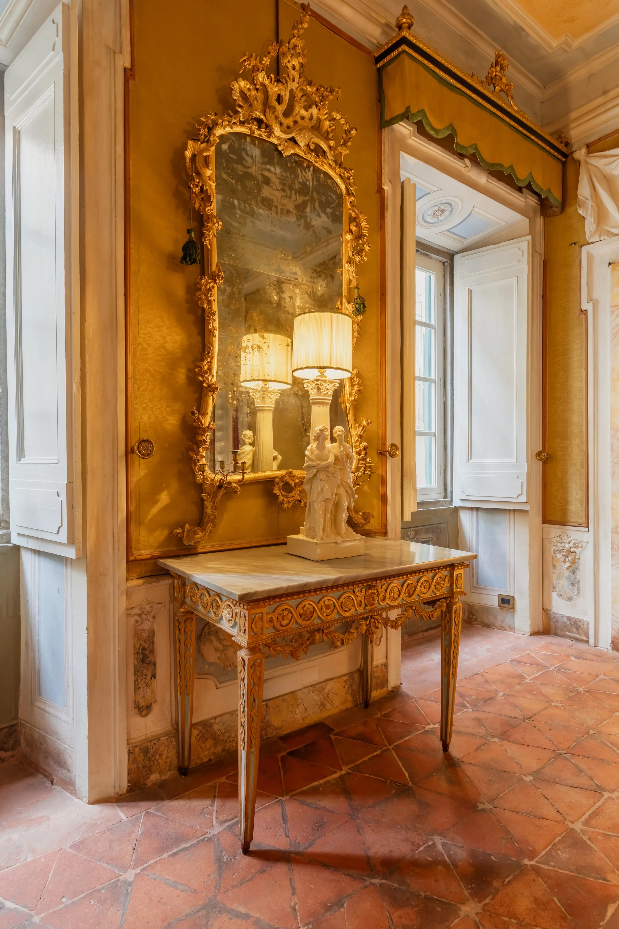 An ornate antique mirror with a gilded, intricate frame leaning against a yellow wall. Below it, a marble-topped table with gold accents holds a white sculpture of a woman holding a child. Two lamps with warm yellow shades illuminate the scene. To th