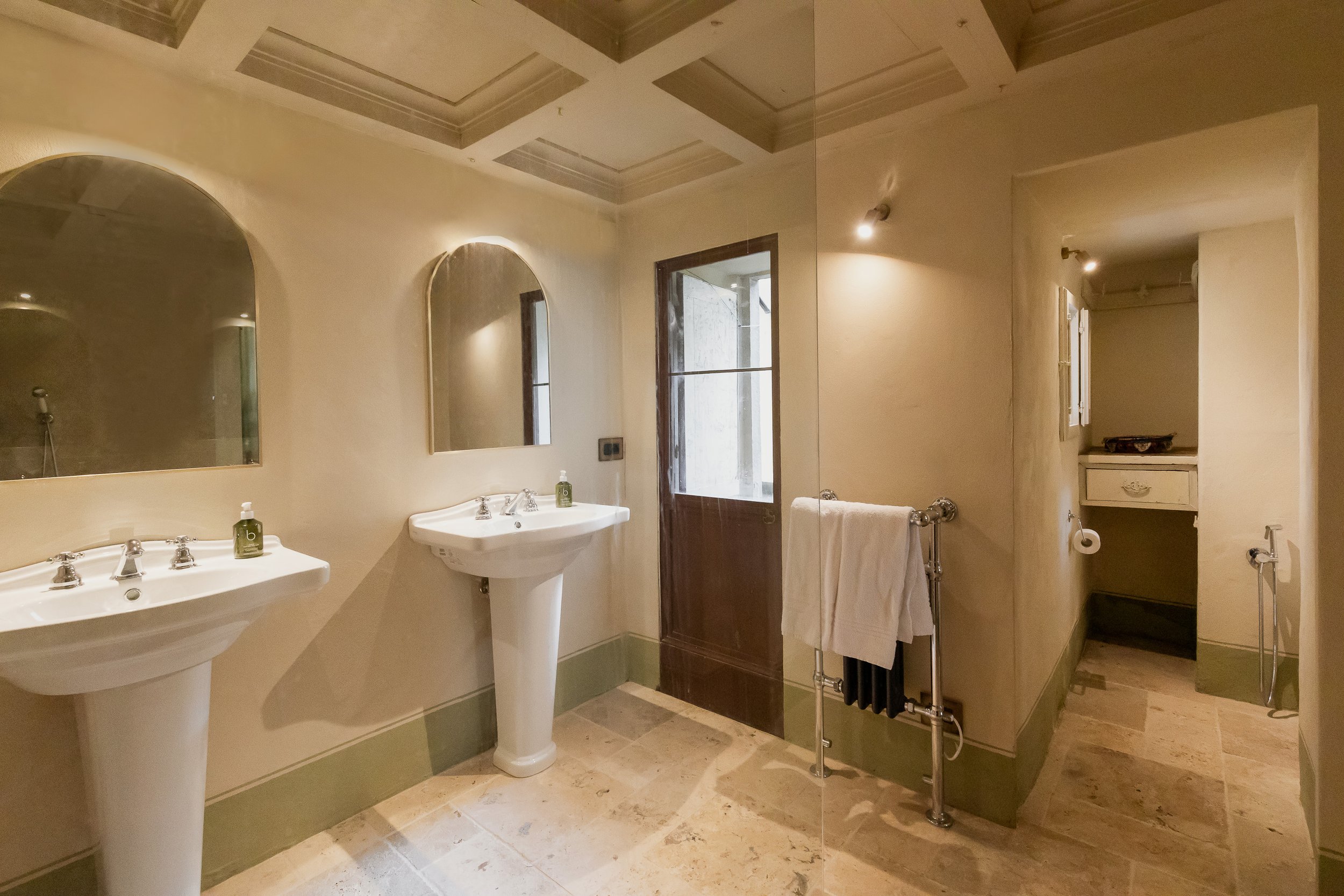 A bathroom with two pedestal sinks, each with a mirror above, a towel rack with towels, a door with a window, and an alcove with a small cabinet and a toilet paper holder. The bathroom has a tiled floor and recessed ceiling lights.