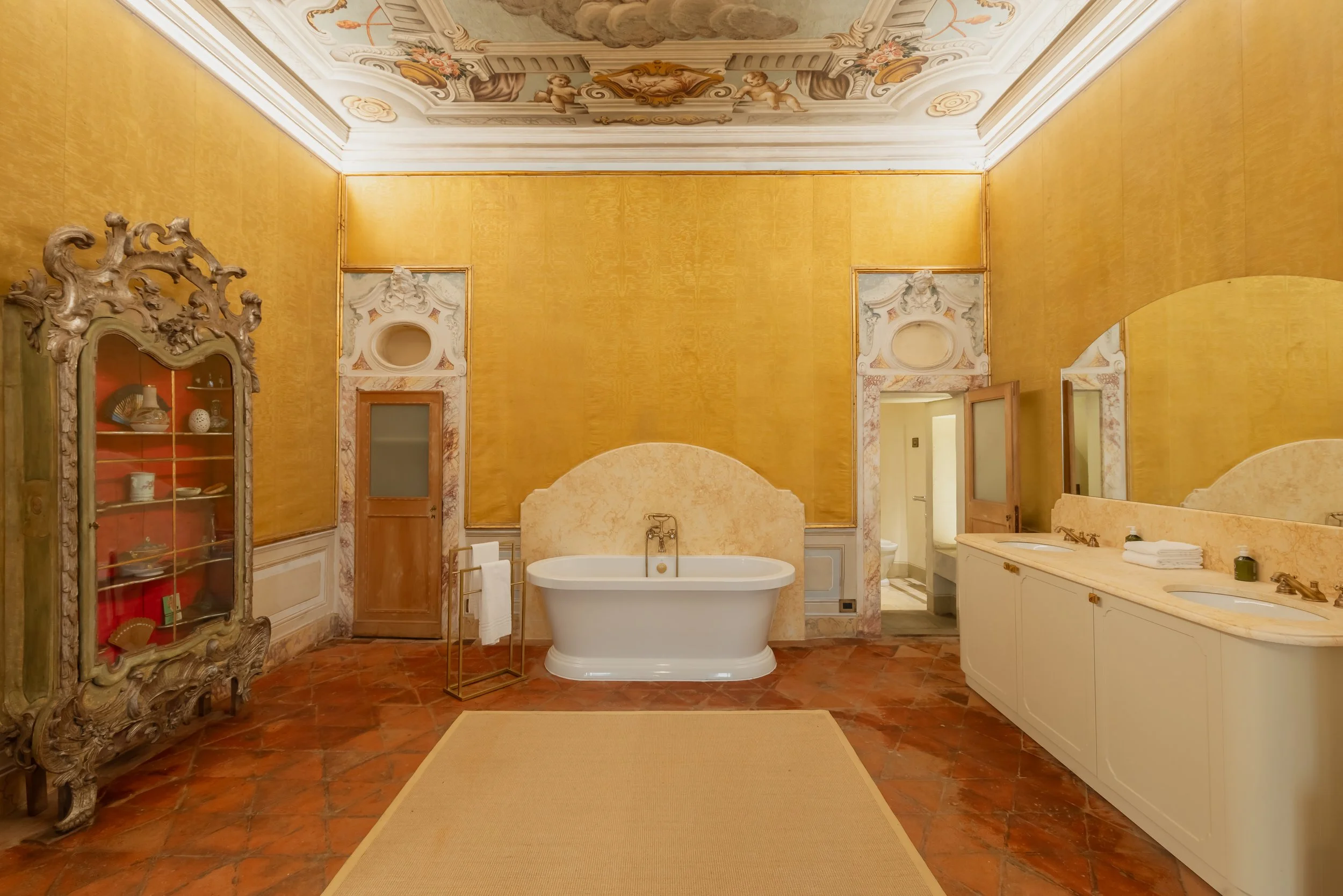 Luxury vintage bathroom with a clawfoot bathtub centered against a yellow wall, ornate cabinet on the left, double sink with mirror on the right, and decorative ceiling artwork.