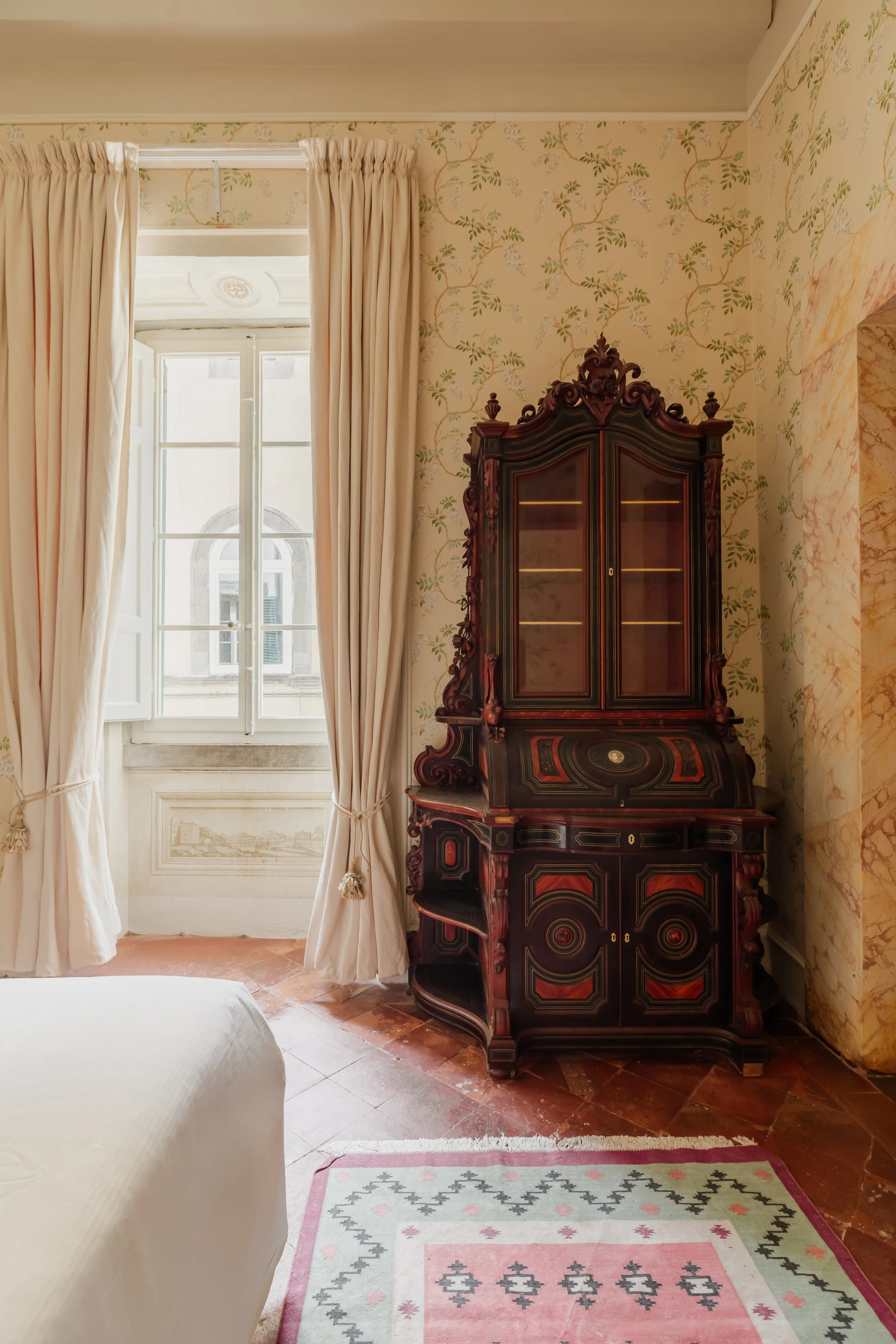 A vintage wooden secretary desk with glass doors in a room with floral wallpaper, a window with cream curtains, and part of a bed and a patterned rug visible.