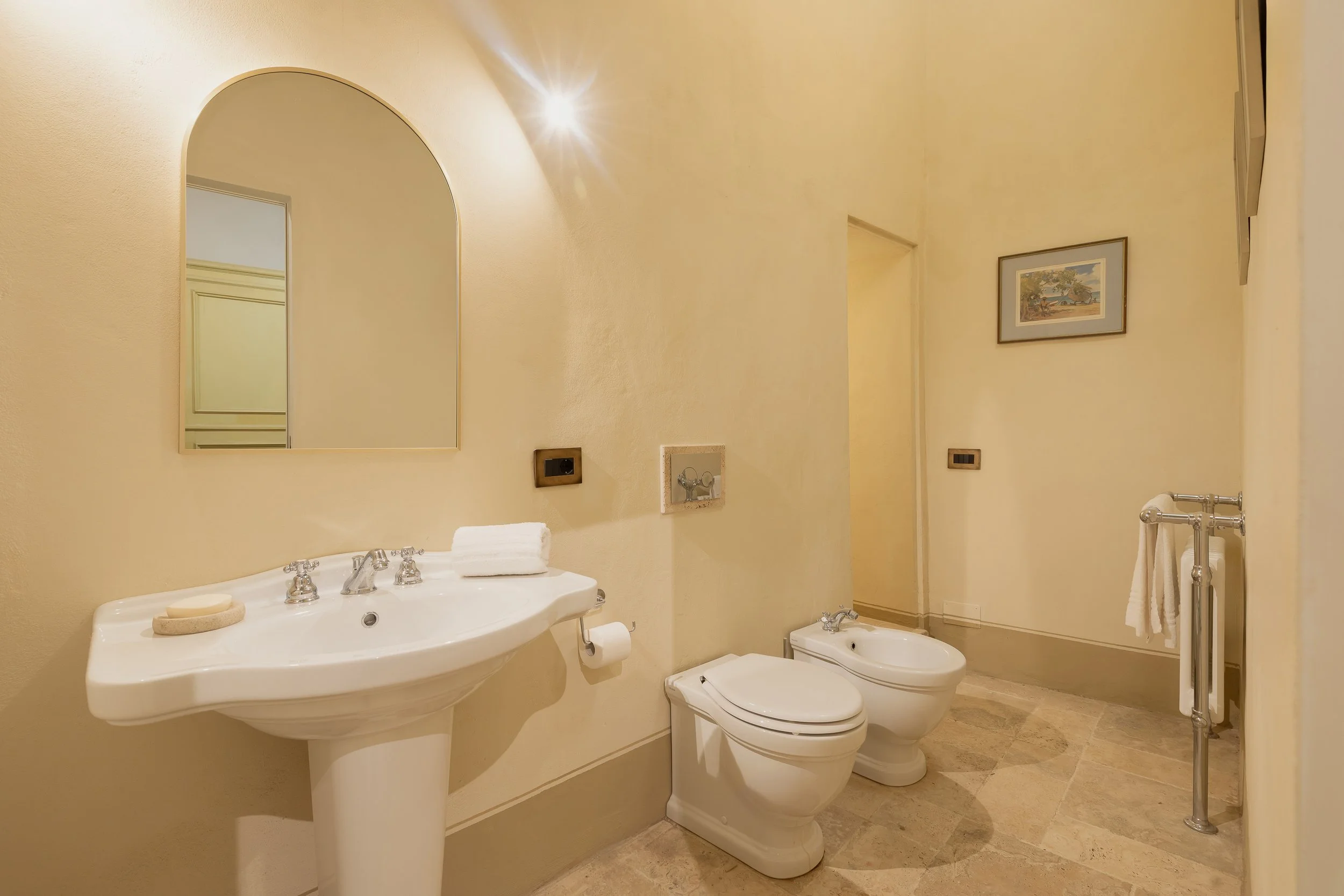Bathroom with a pedestal sink, mirror, toilet, bidet, towel rack, and framed artwork on cream-colored walls.