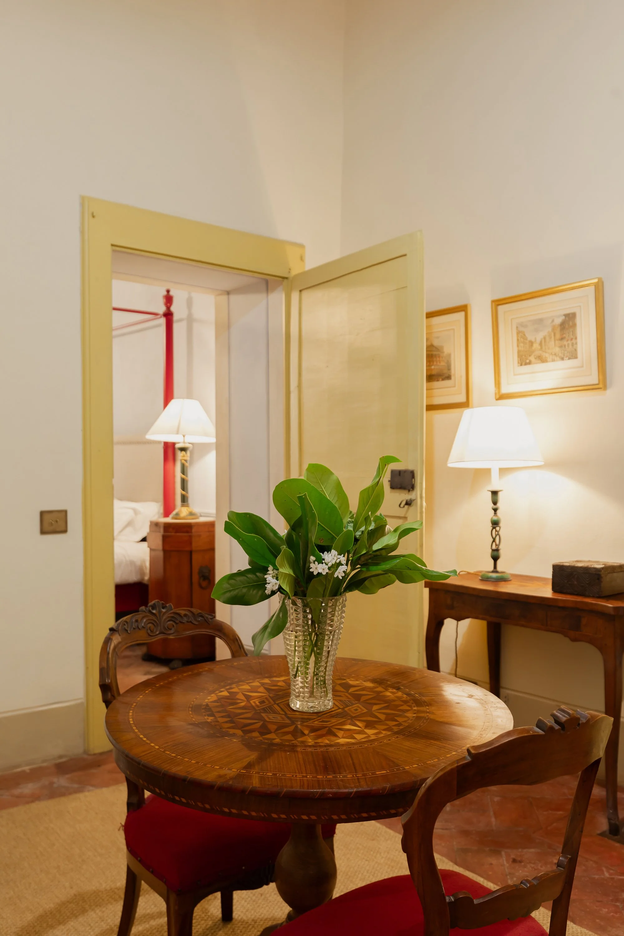 A round wooden dining table with a glass vase containing green leafy plants and white flowers. Surrounding the table are three wooden chairs with red upholstered seats. In the background, there is a side table with a lamp and two framed pictures on t