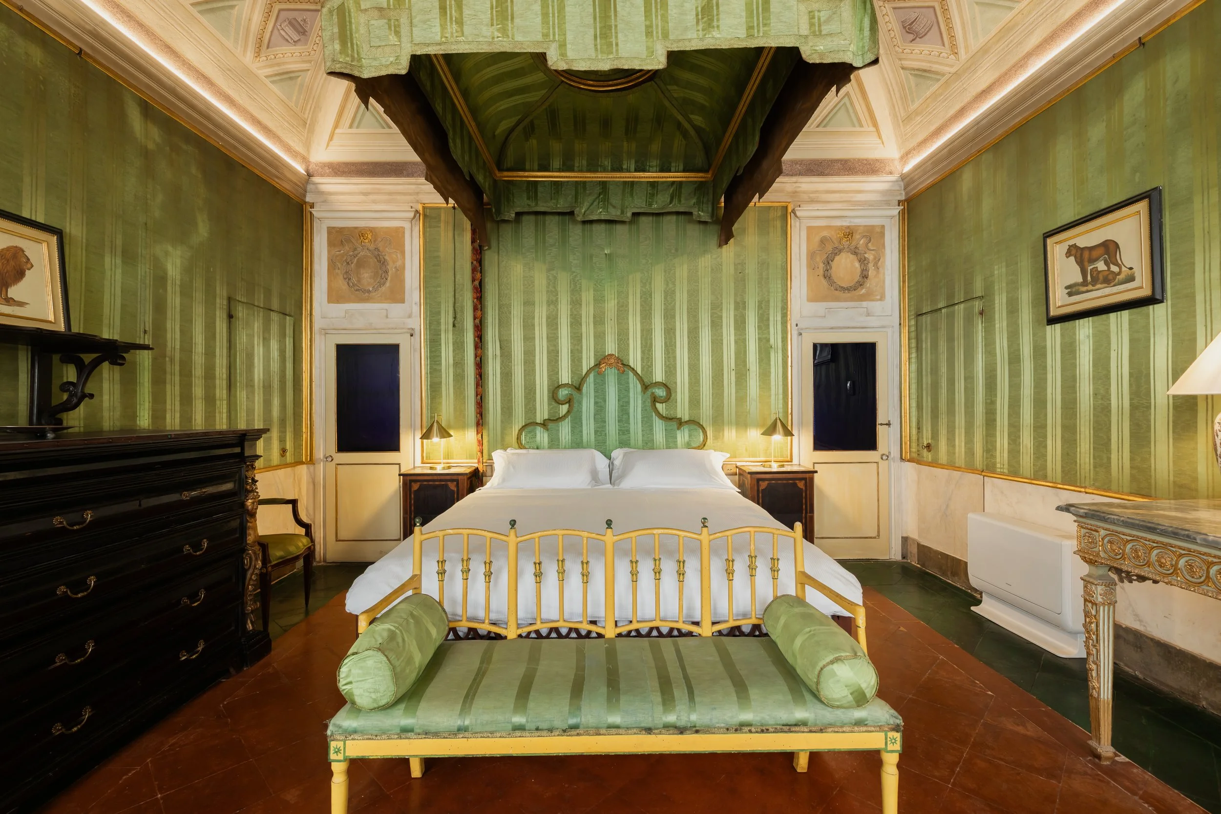A luxurious bedroom with green striped wallpaper, a yellow metal bed with white bedding, and matching green cylindrical pillows at the foot of the bed. There are two nightstands with lamps on either side of the bed, and a black dresser with gold hand