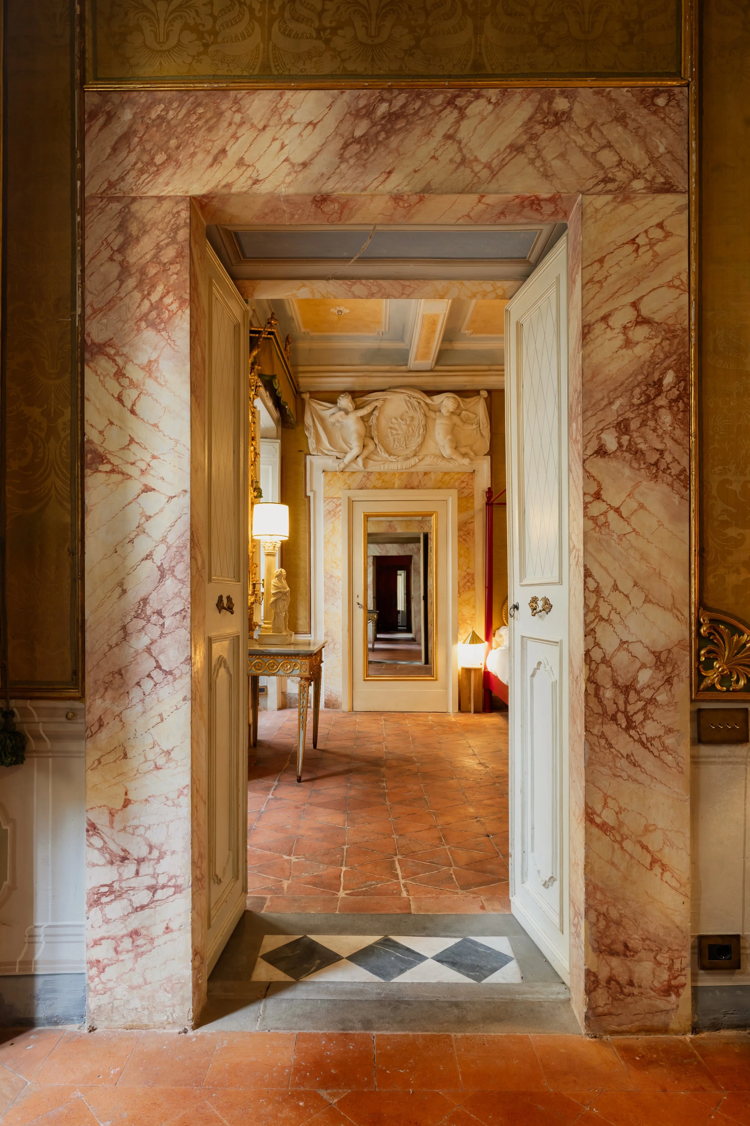 Interior view of a luxurious, ornately decorated room with marble and gold accents, including a doorway leading to another elegant room with a tiled floor and classical statues.