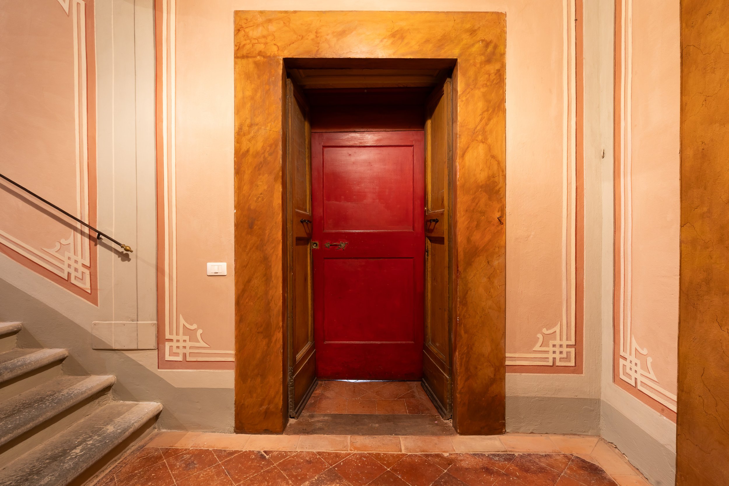 An interior scene showing a red door with historical decor, framed by two golden-brown marble or faux-marble walls, and a staircase on the left with stone steps and a black handrail.