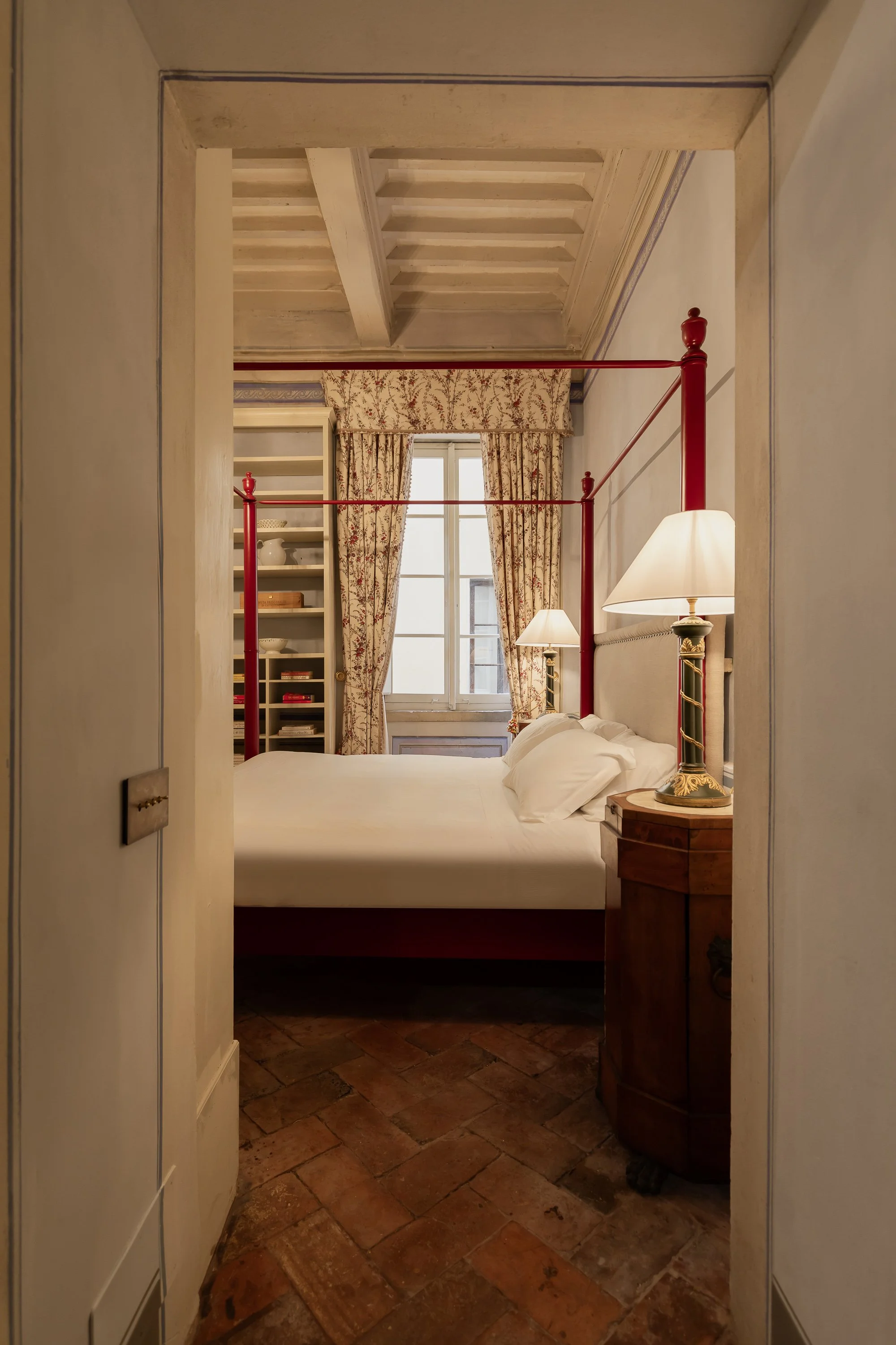 A view of a hotel room featuring a four-poster bed with white linens, wooden side table with a decorative lamp, a window with floral curtains, and a built-in bookshelf.