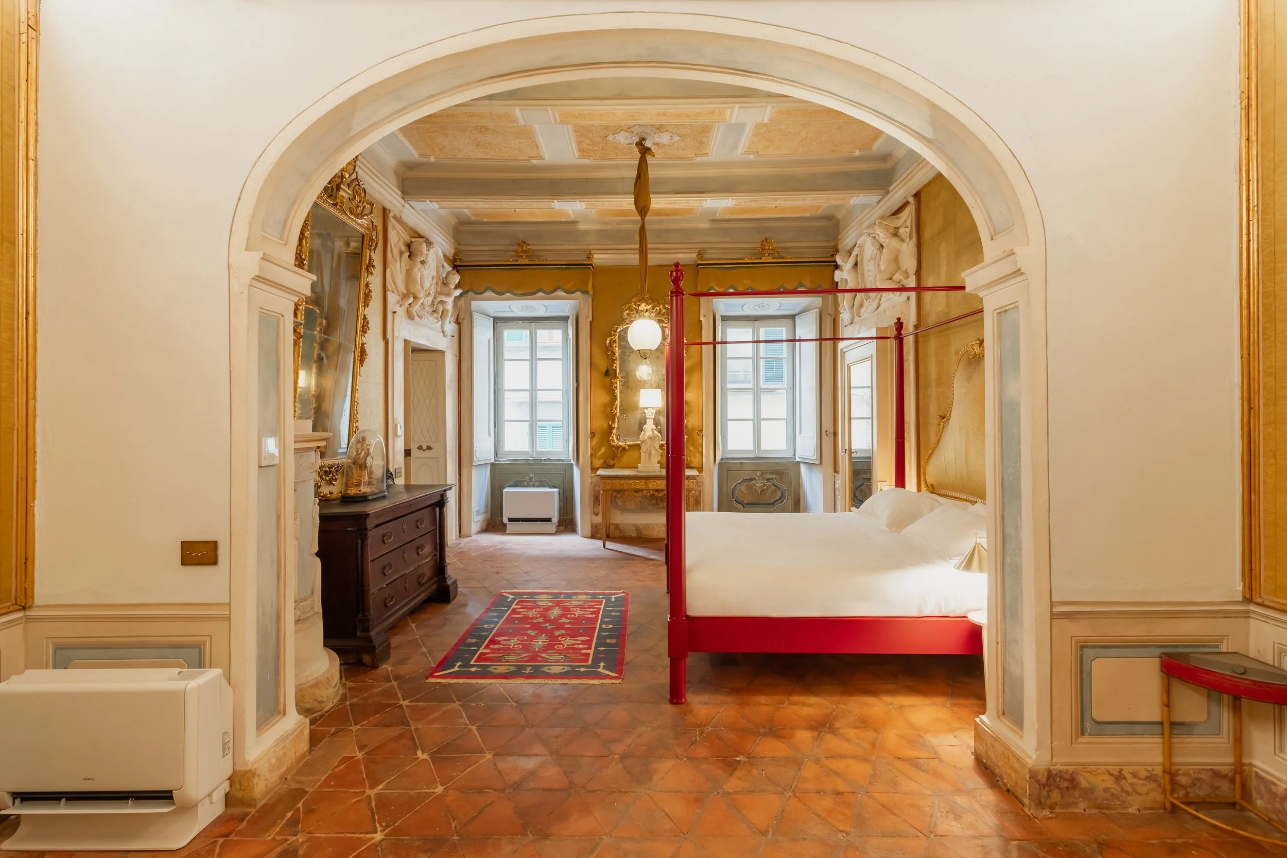 A luxurious bedroom with ornate decor, a red four-poster bed, large windows with shutters, antique furniture, and decorative wall sculptures.