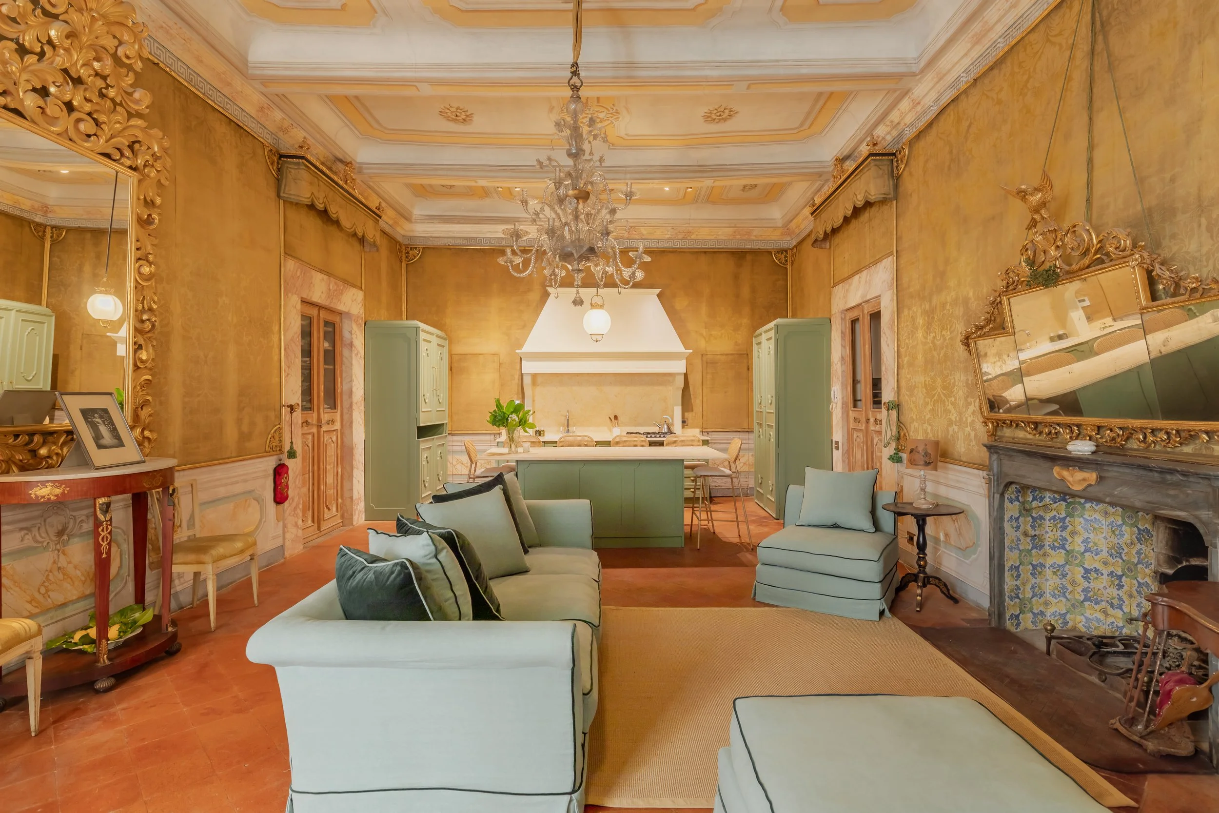 A luxurious living room with gold patterned walls, a large ornate chandelier on the ceiling, a fireplace with decorative details, a mirror, green cabinets, and light-colored couches with cushions.
