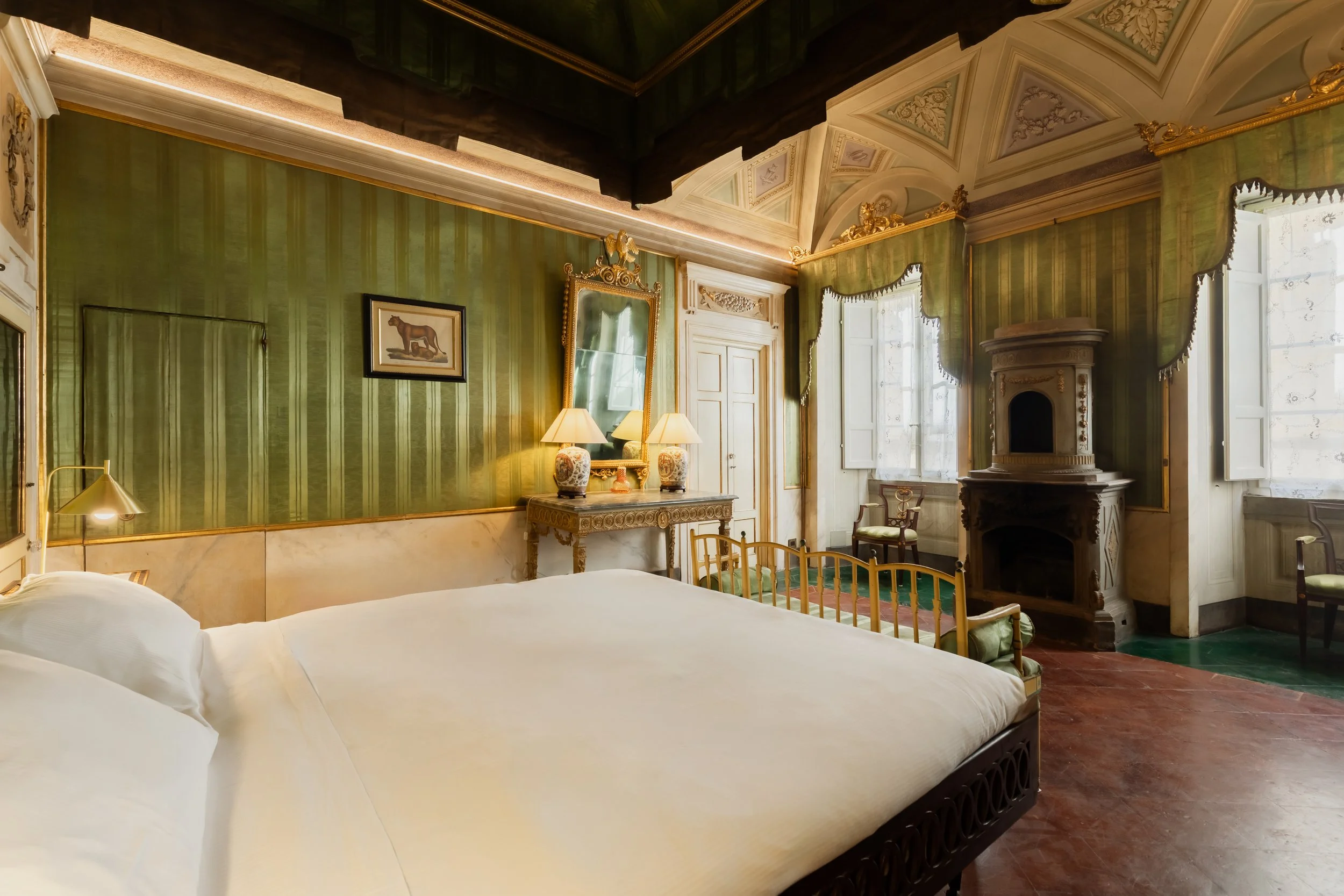 Elegant bedroom with green striped walls, white bedding, antique furniture, a fireplace, and large windows with drapes.